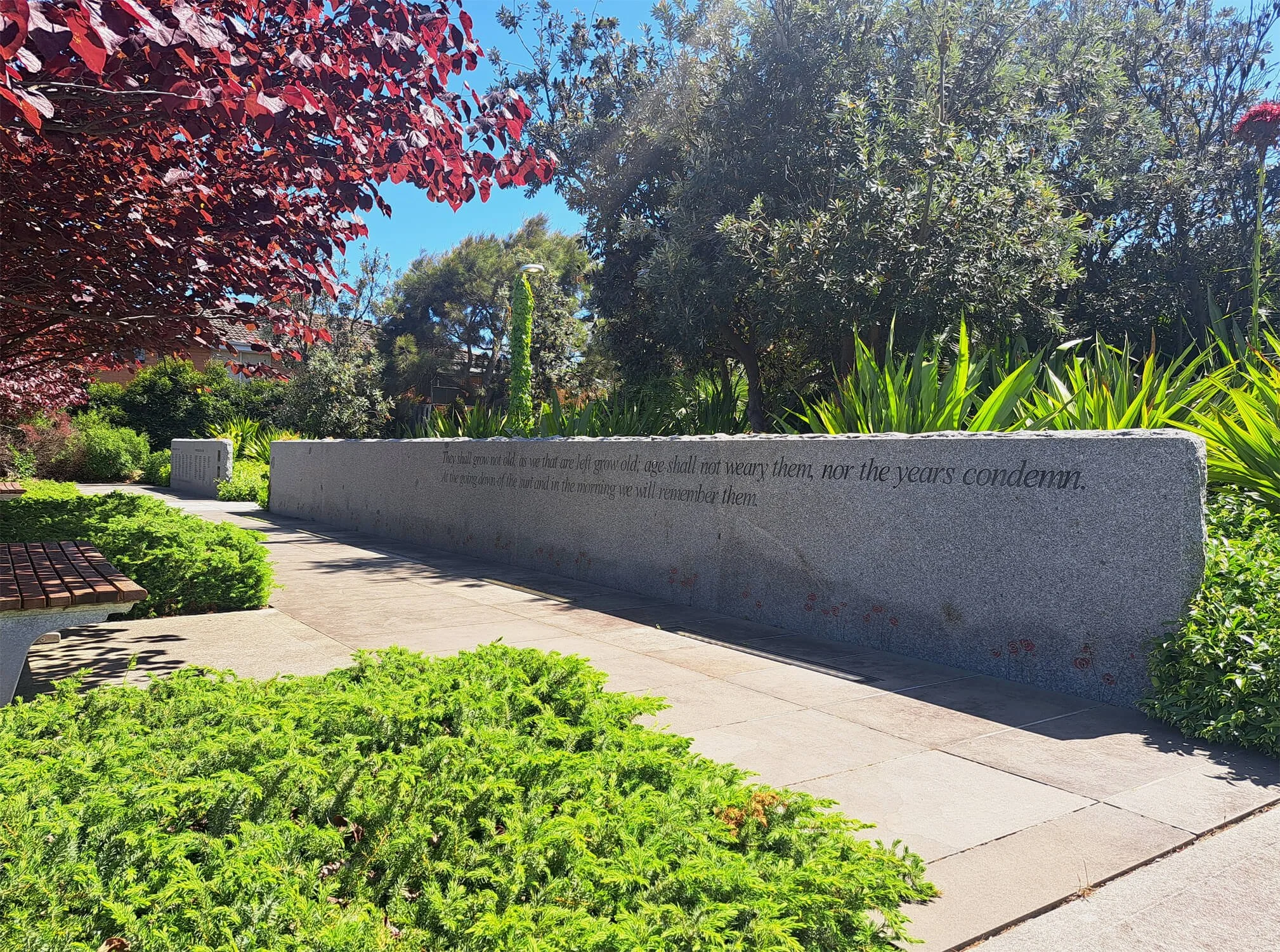 A garden with various green plants and trees, a stone bench, and a long stone wall with engraved writing, under a clear blue sky.