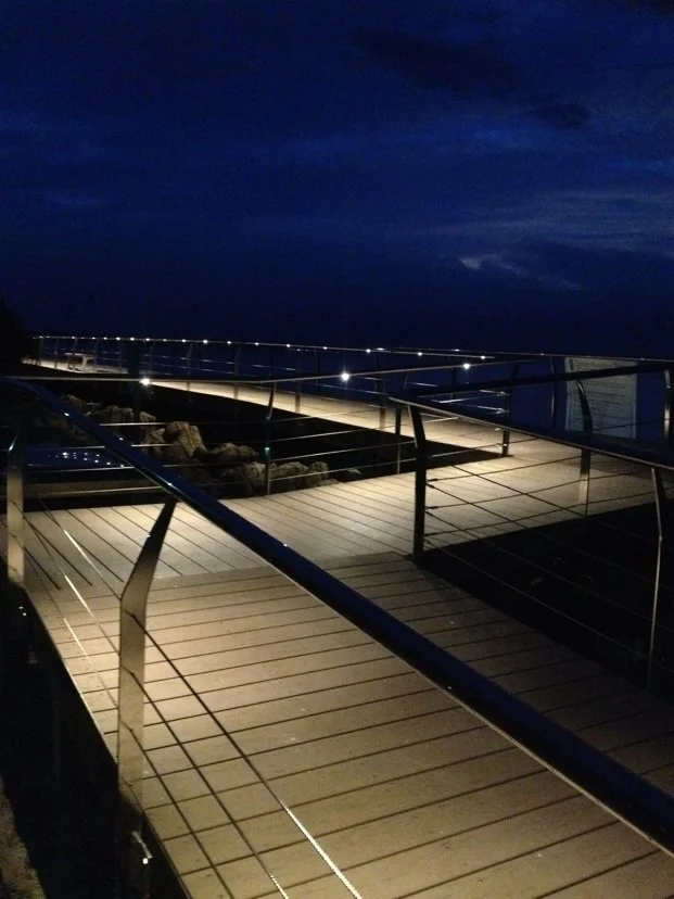 Empty wooden boardwalk with metal railings at night, overlooking a body of water with dark skies and some clouds.