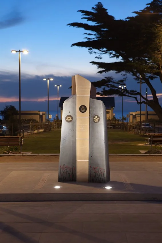 A memorial monument with three plaques, illuminated by ground lights, set in a park during evening, with trees and buildings in the background.