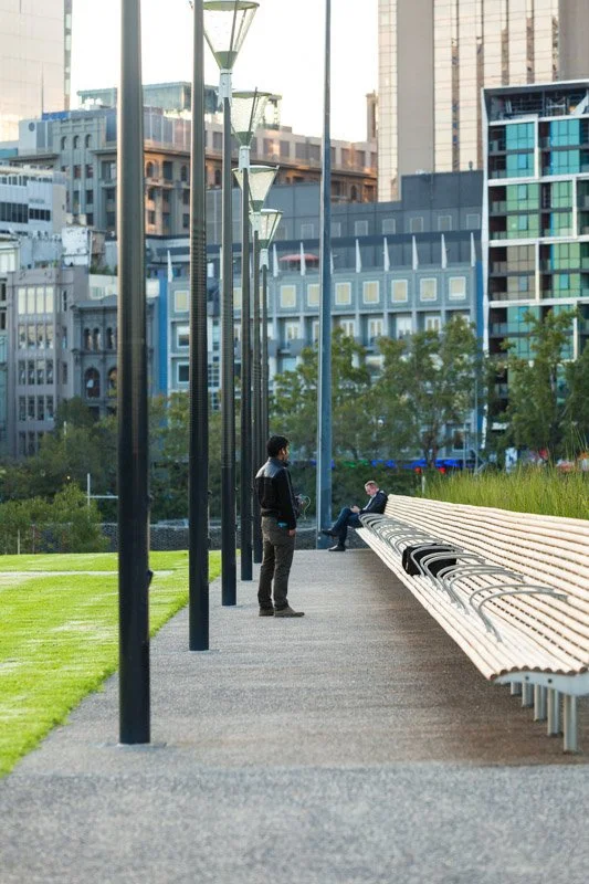 Two people in a city park: one standing near a row of street lamps, and another sitting on a long bench with a grid-like pattern, surrounded by greenery and tall buildings in the background.