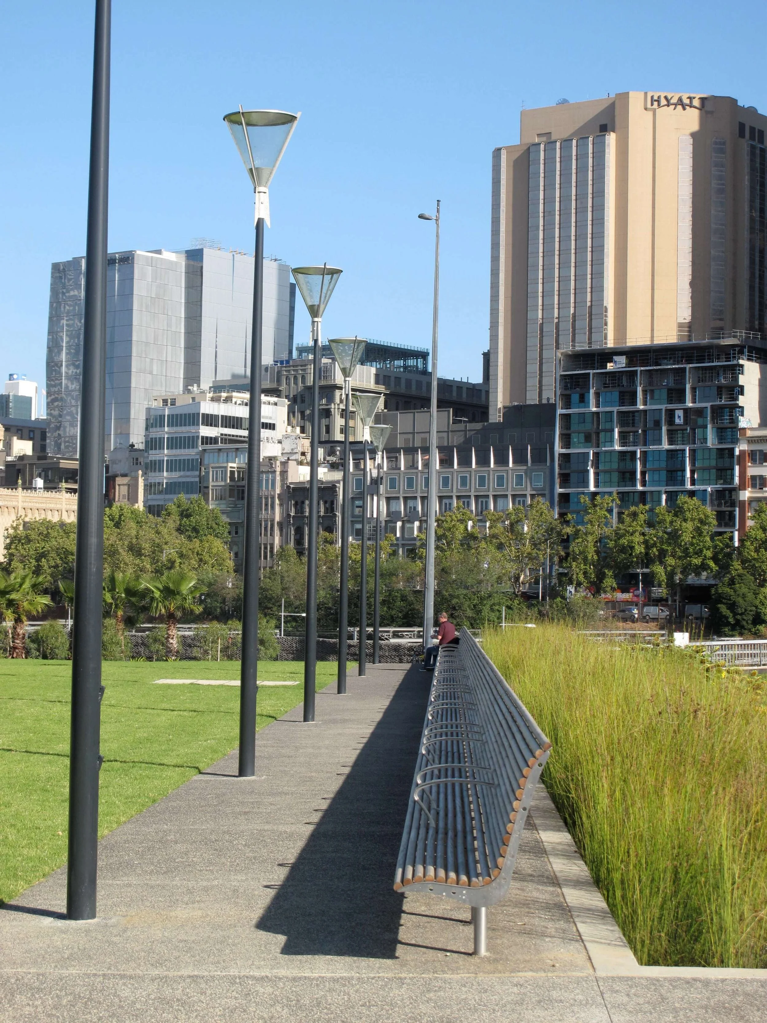 A city park with a walking path, modern street lamps, a bench, and buildings in the background, including a hotel labeled Hyatt.