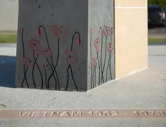 Graffiti of pink flowers with black stems and leaves drawn on the corner of a concrete and brick structure, with a carved inscription reading 'Vietnam 1962 - 1975' on the ground nearby.