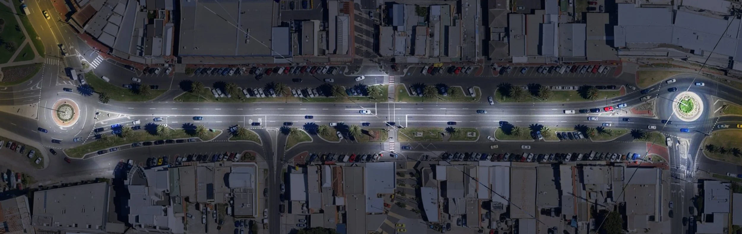 Aerial view of a city street at night with cars, streetlights, and small green spaces with palm trees and circular traffic islands.