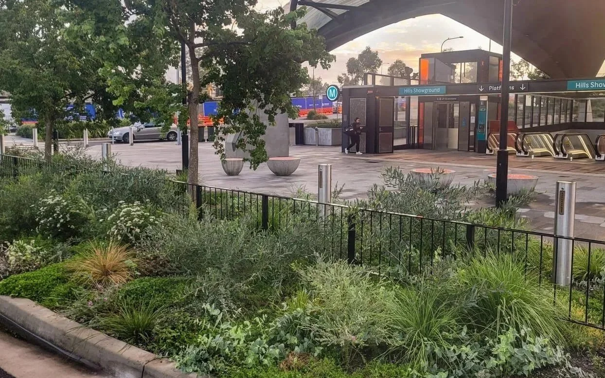 Entrance to the Hills Showground subway station with escalators leading inside, surrounded by greenery and trees, parked cars, and a person sitting on a bench.