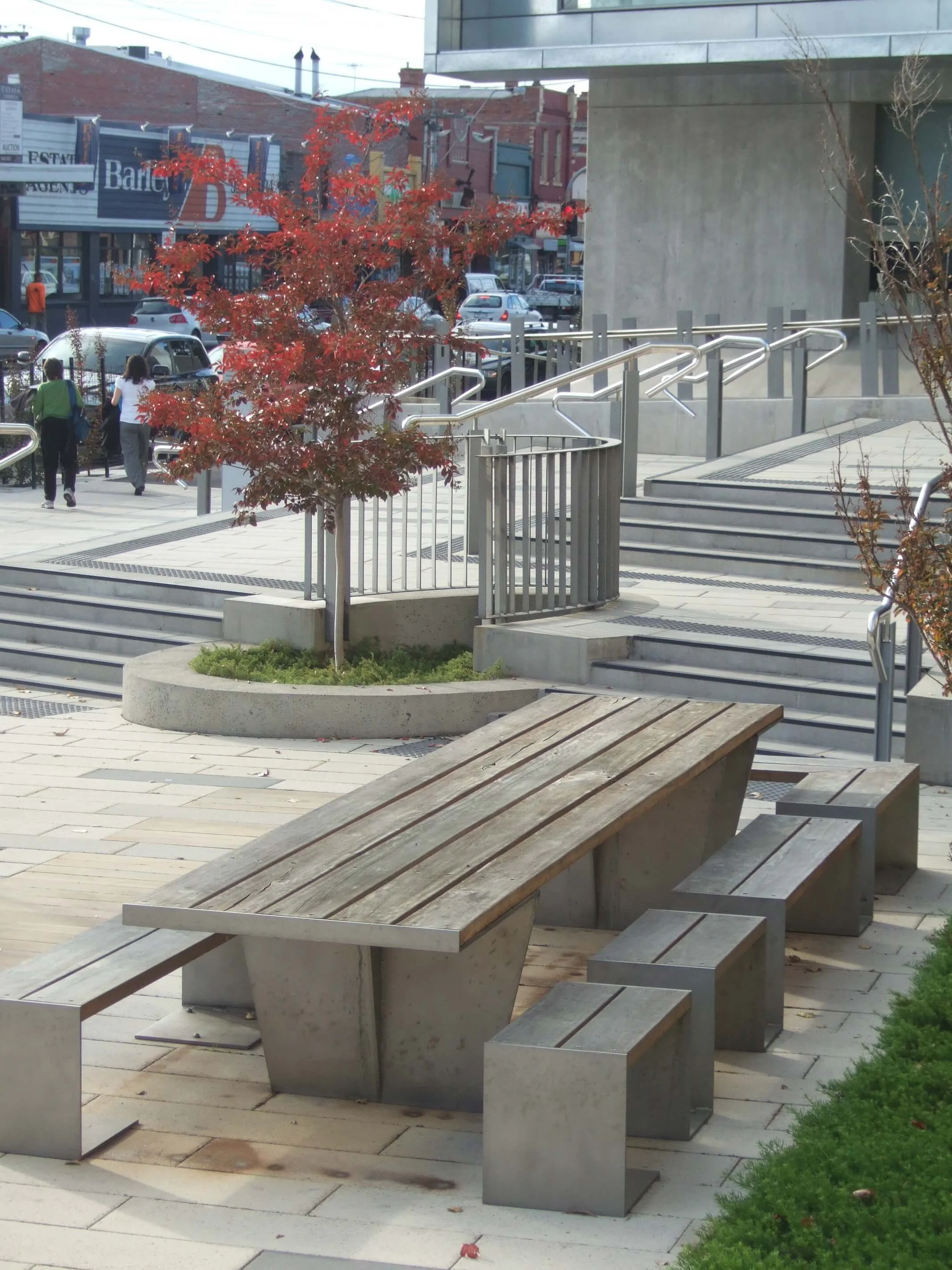 Outdoor urban scene with a wooden-and-concrete bench, small trees with red leaves, stairs with metal handrails, and a few pedestrians walking in the background.