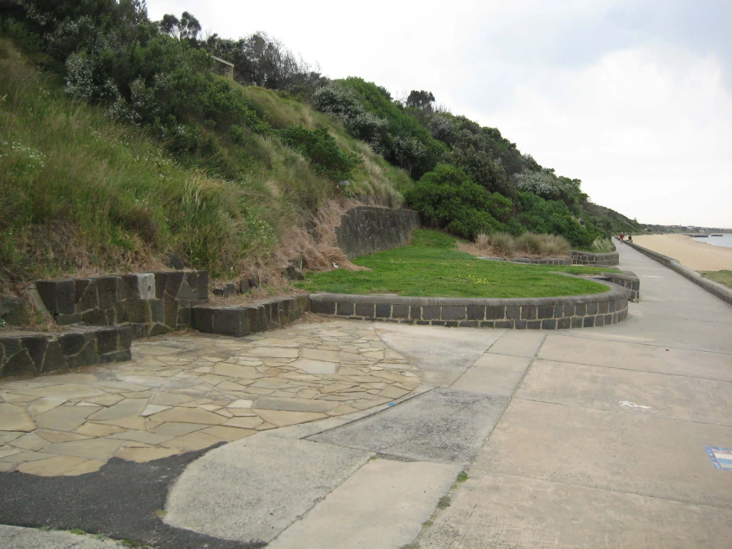 A coastal walkway with a stone and concrete path, a grassy area, and a hillside covered with green bushes and some trees, extending toward the sandy beach and ocean in the distance.