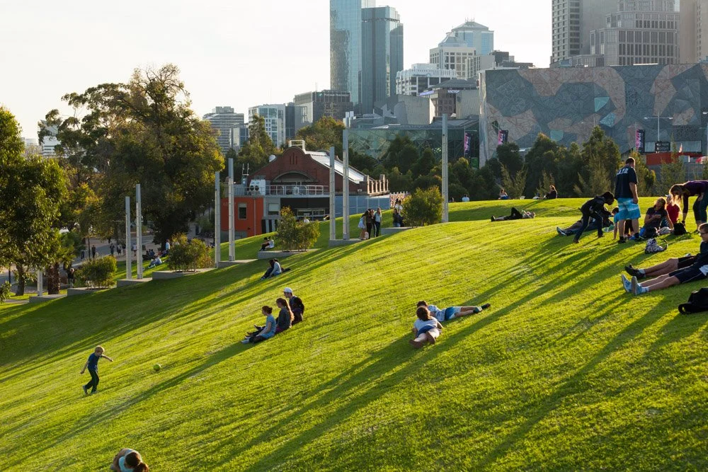 People relaxing and playing on a grassy hill in an urban park with city skyscrapers in the background during sunset.