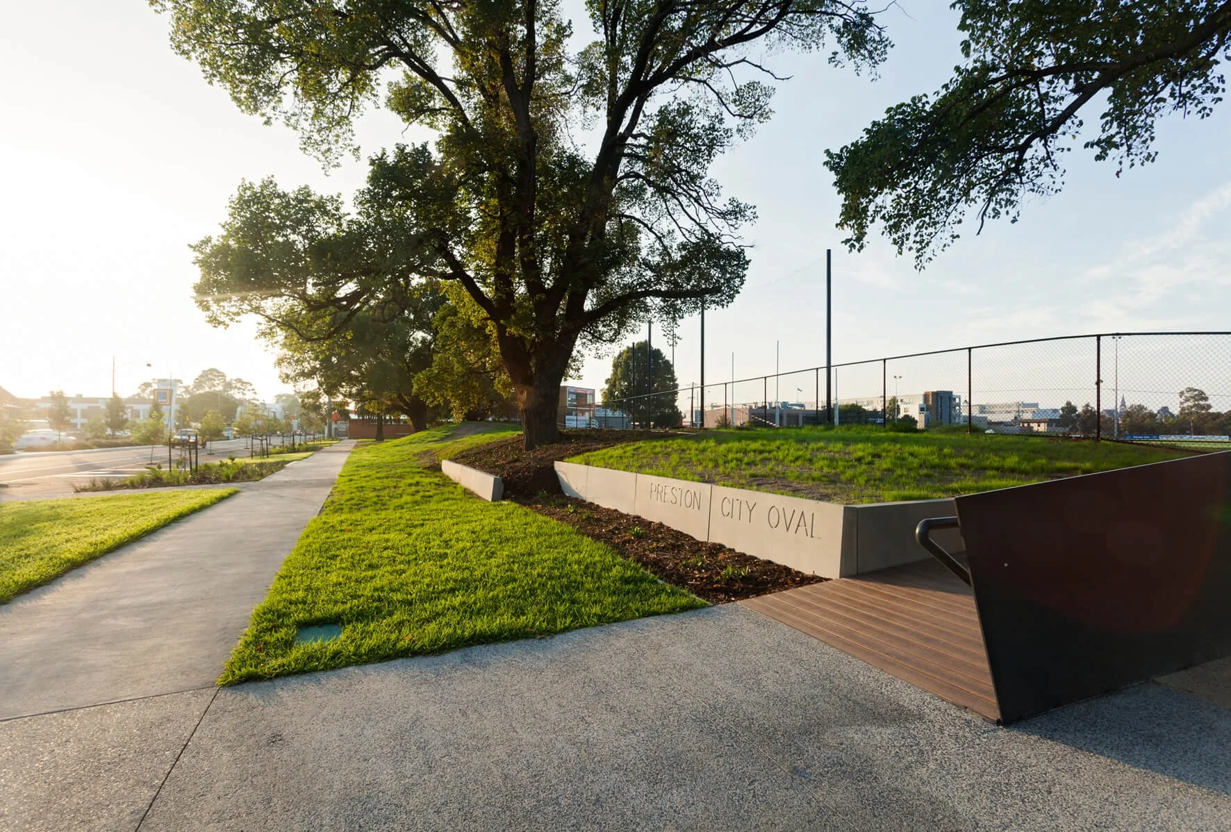 Sidewalk and grassy area with a large tree, a bench, and a fence at Presiton City Oval.