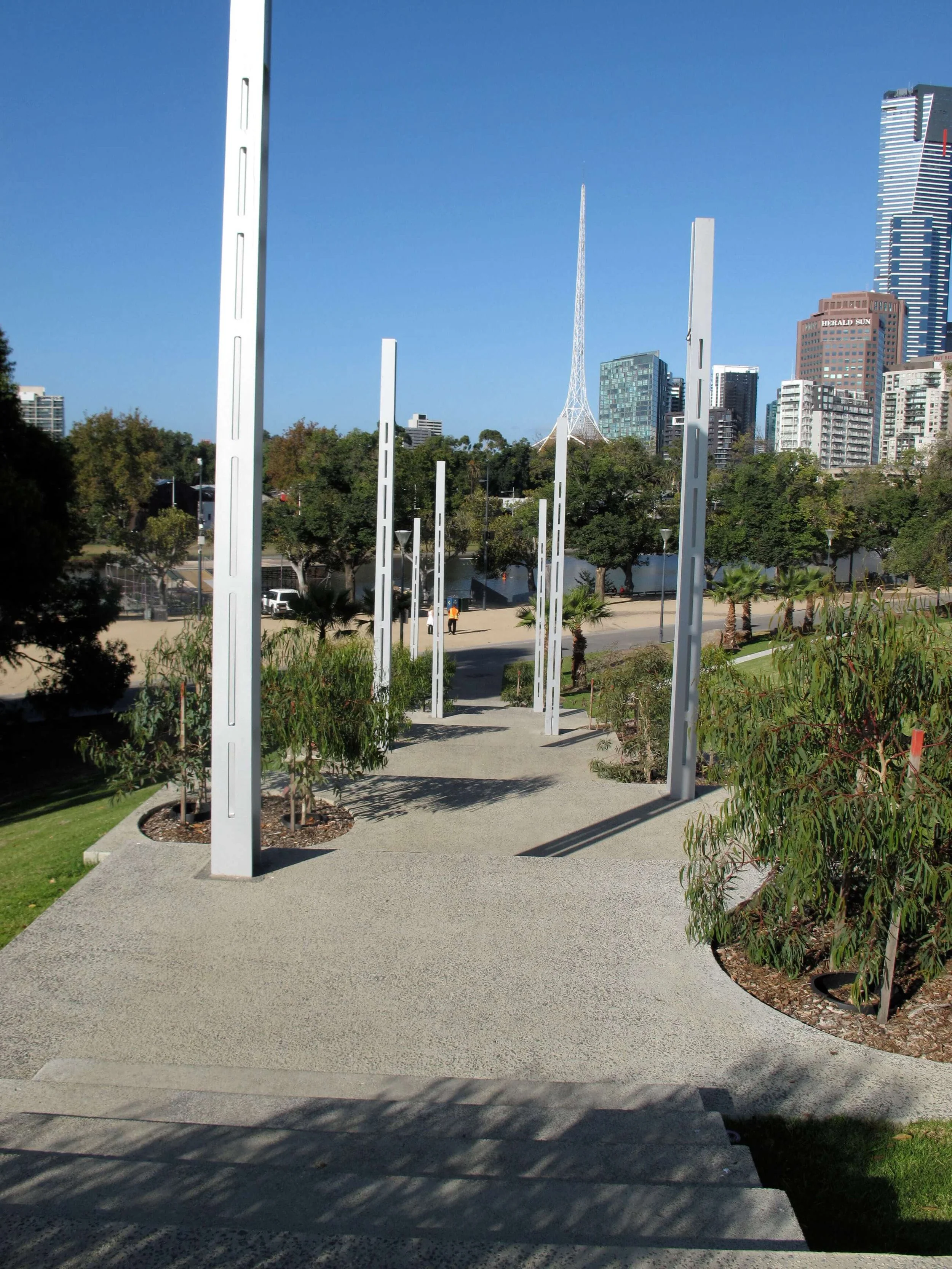 City park with modern white sculptures lining a pathway, trees and greenery in the background, and tall city buildings under a blue sky.