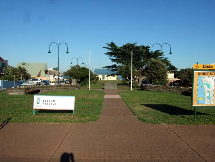 A small park with a brick walkway, two benches, and a large tree. Signage indicates it is Beeson Reserve, with street lamps and buildings in the background.