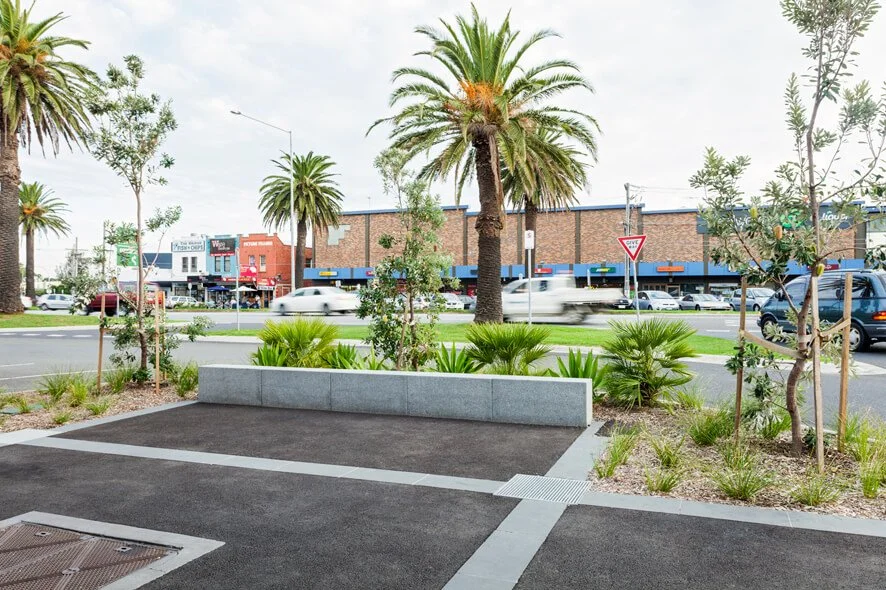 View of a landscaped public space with a small parking area, surrounded by palm trees and other greenery, with a busy street and commercial buildings in the background.