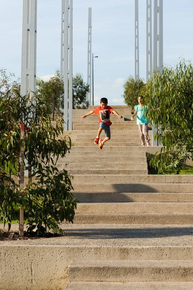 Two children, a boy and a girl, playing on outdoor concrete steps, with the boy jumping and the girl holding his hand, surrounded by greenery and modern metallic structures.