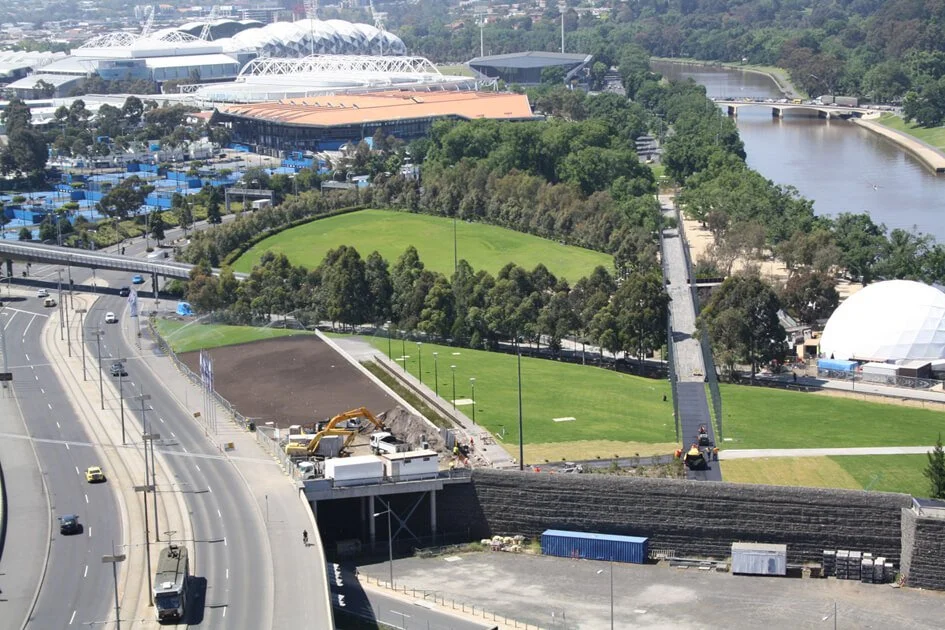 Construction work on a bridge over a highway near a park with green lawns, trees, and a river, with sports stadiums in the background.