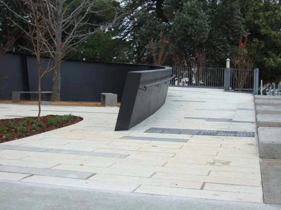 Public outdoor space with paved walkway, curved black metal structure, a tree, and a railing in the background.