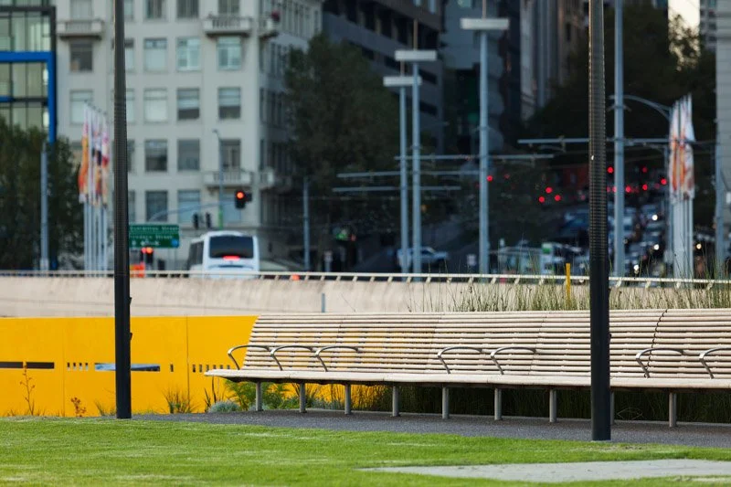 View of an urban park with wooden benches, grass, and pathways, with city buildings and traffic lights in the background.