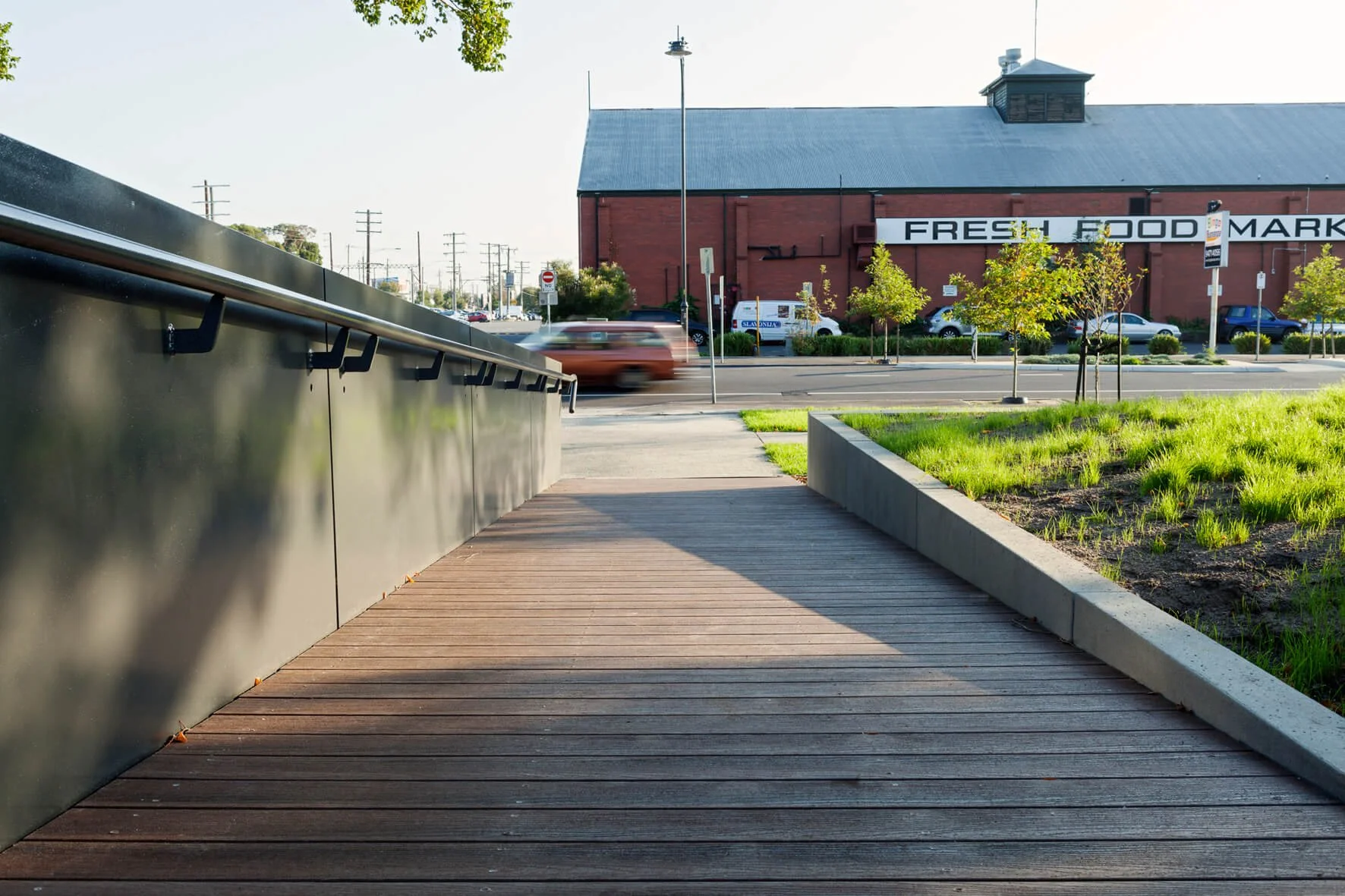 A wooden sidewalk with a metal railing on the left, a grassy area with young trees on the right, and a red brick building with a sign that reads 'FRESH FOOD MARKET' across the street. Cars are passing by on the street in front of the building.