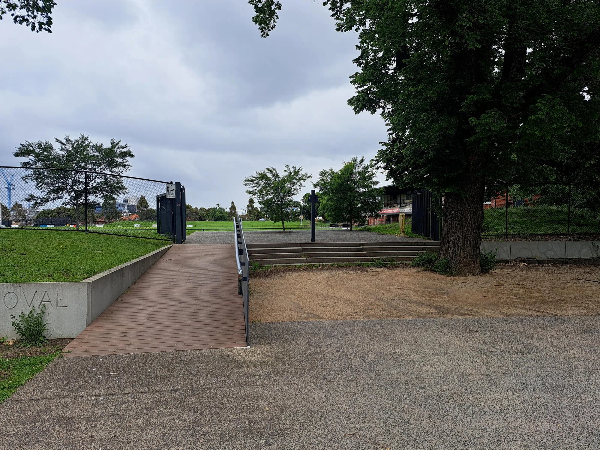 View of a park entrance with a concrete and wooden ramp, steps, a tree, grass, a sidewalk, chain-link fence, and some buildings and trees in the distance under an overcast sky.