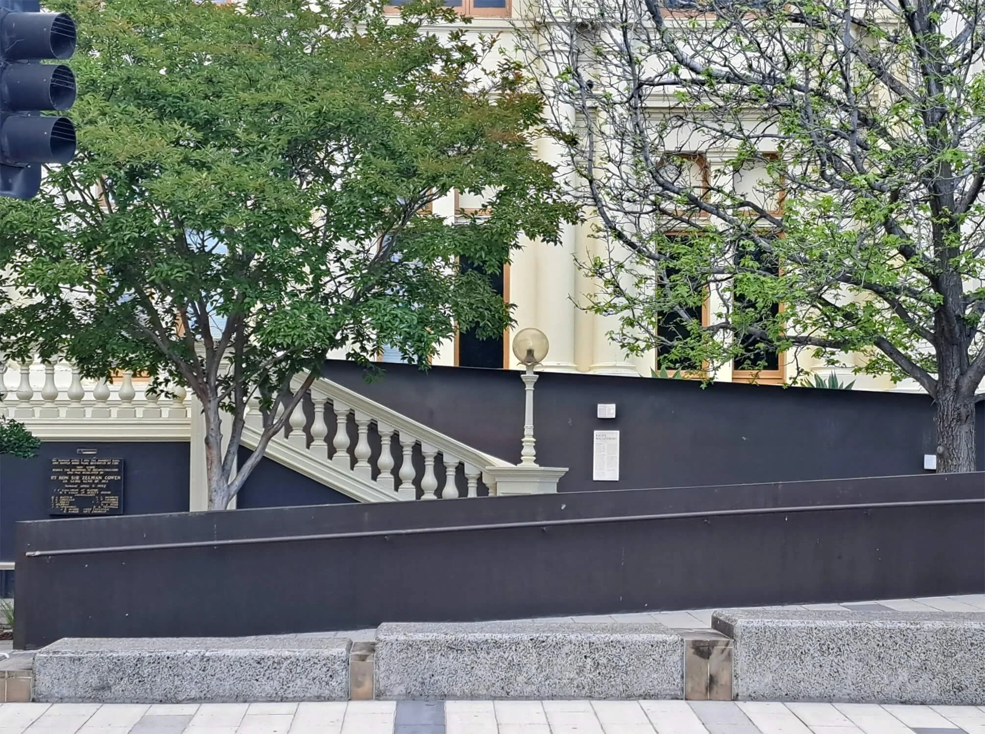 Street view of a building with a staircase leading up to the entrance, flanked by trees and a black wall, with a streetlamp on top of the wall.