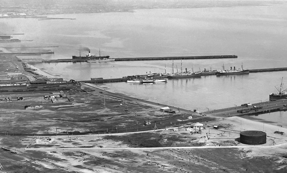 Aerial view of a large harbor with ships docked, industrial buildings, and a vast storage tank, under a cloudy sky.