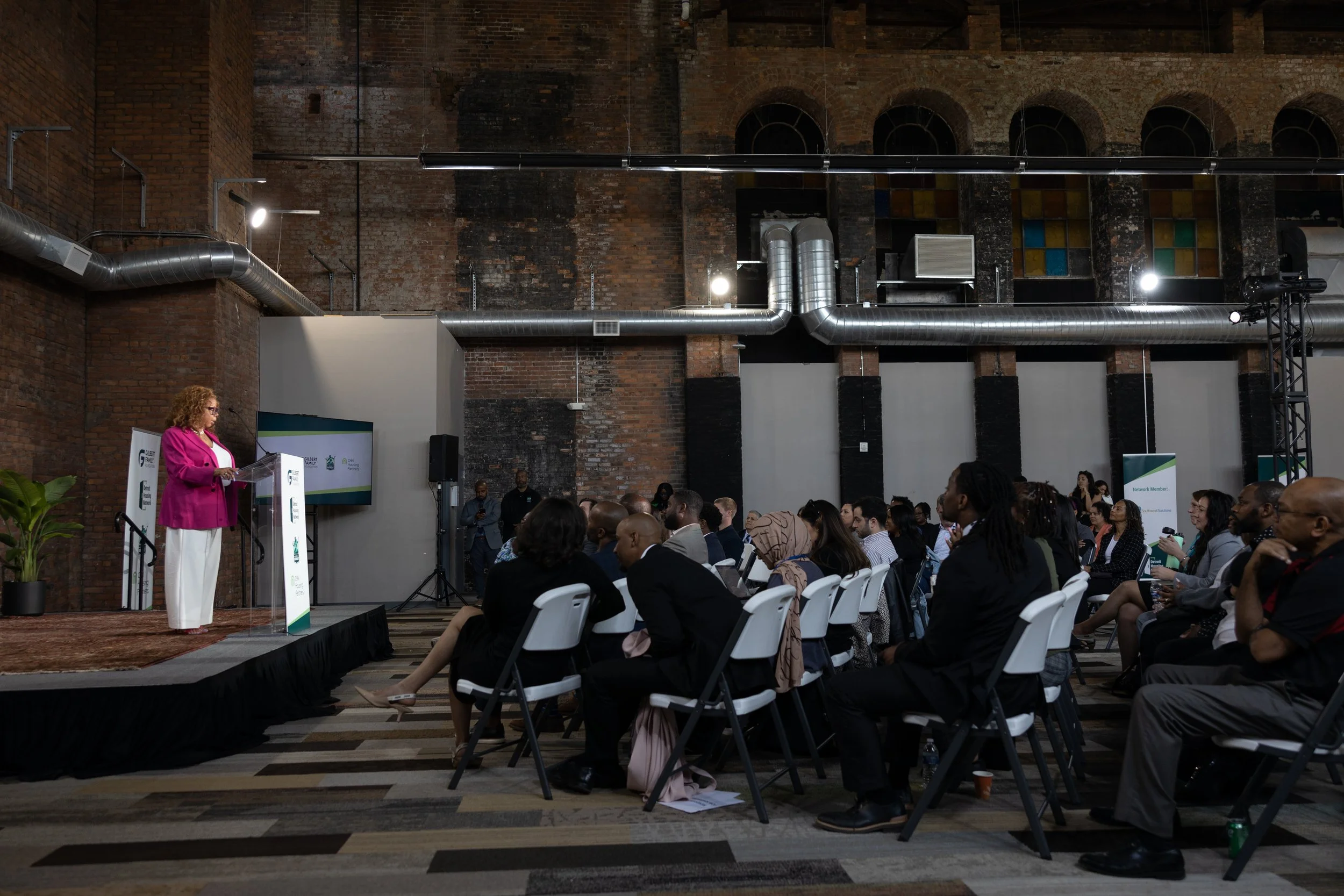 A woman in a pink blazer and white pants standing at a podium, speaking to an audience seated in white chairs in an industrial-style conference room with exposed brick walls and ductwork.