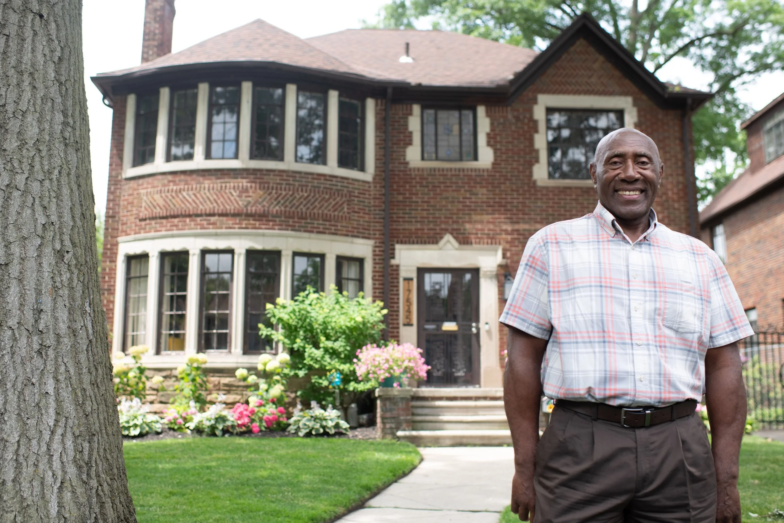 A smiling man stands in front of a large brick house with a well-maintained garden.
