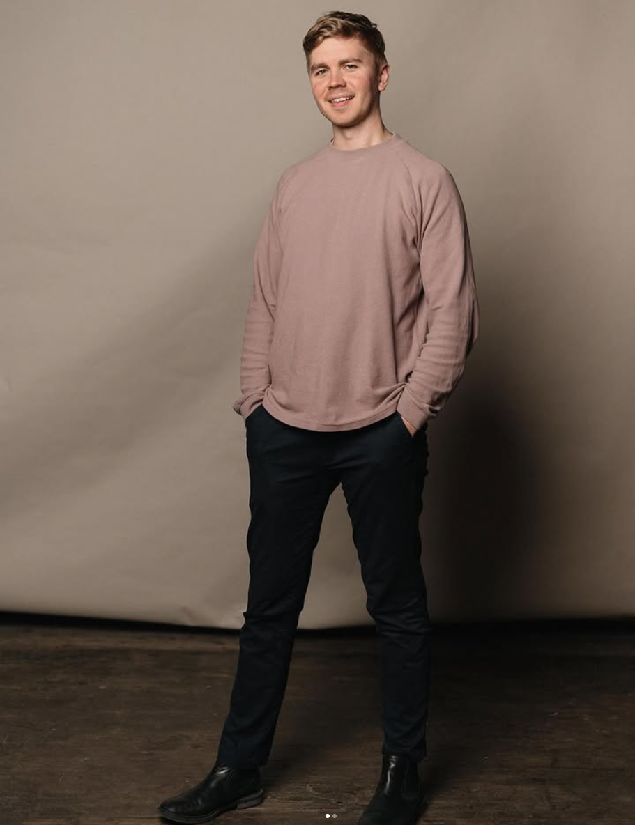 A young man with short, wavy brown hair standing in front of a plain beige background. He's smiling slightly, wearing a light pink crewneck sweatshirt, dark pants, and black boots, with hands in his pockets.