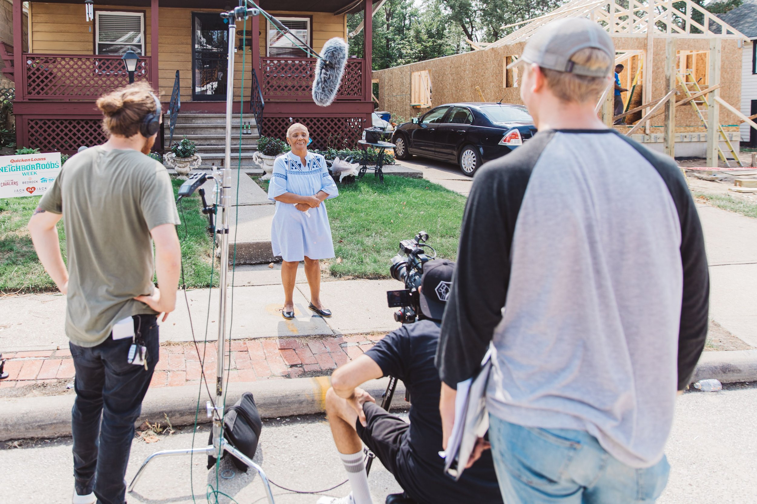 A film crew is filming a woman speaking outside a house under construction. The woman is standing on the sidewalk, smiling, wearing a blue and white dress. The crew includes a camera operator, a person with headphones, and another person with a clipboard, with a microphone boom overhead.