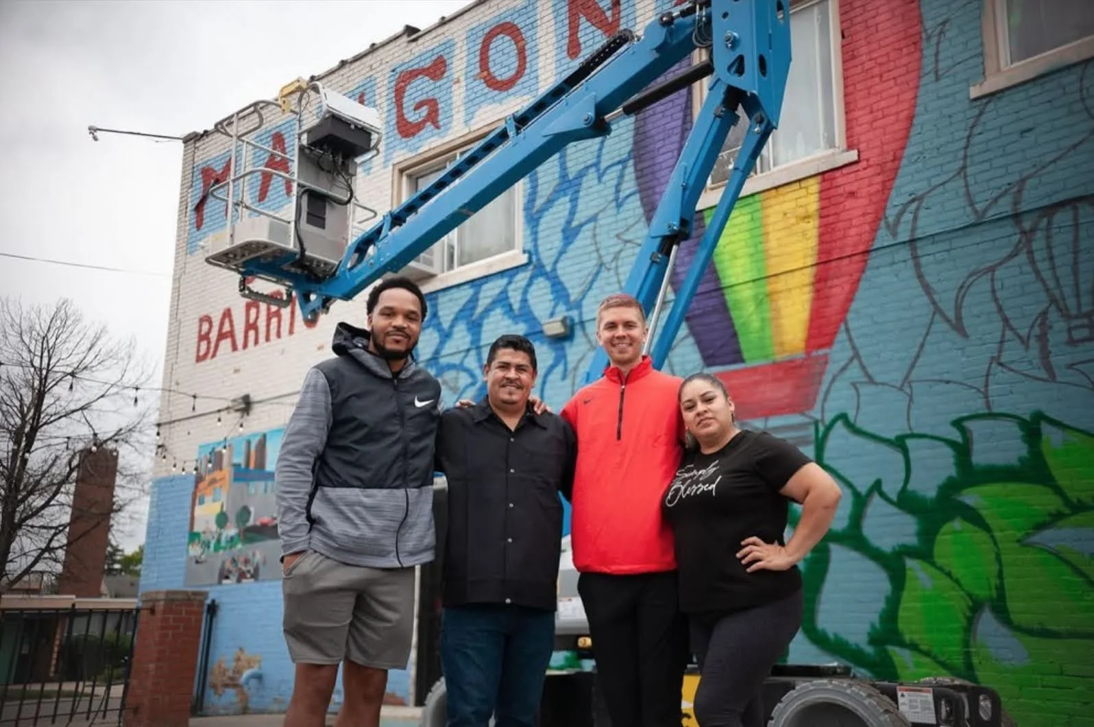 Four people standing in front of a colorful mural on a brick building, some on a lift or bucket truck.