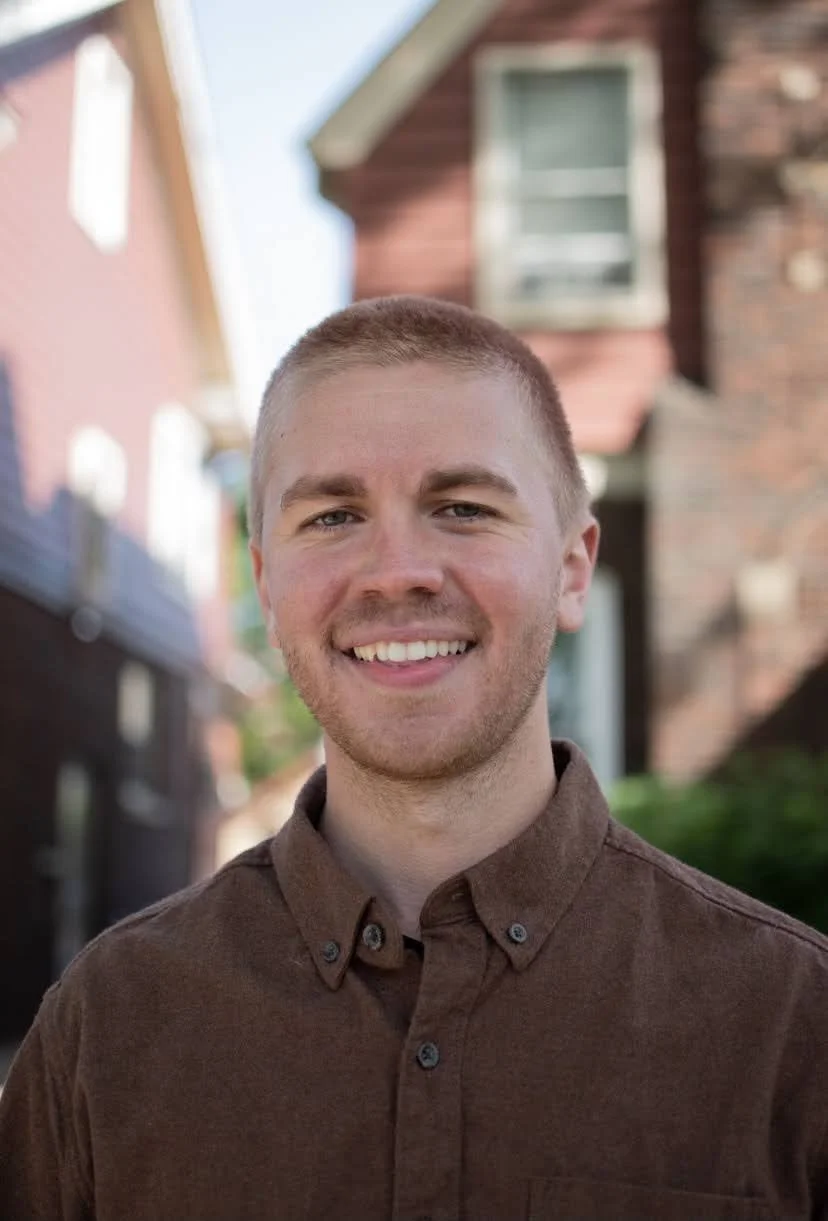 A young man smiling outdoors in front of houses with colorful facades, wearing a brown button-up shirt.