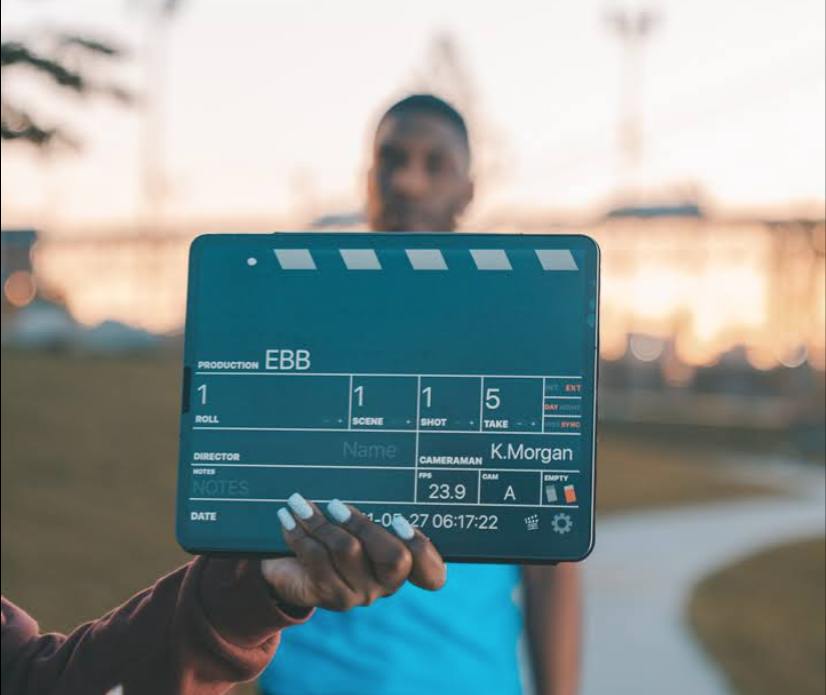 A person holding a film clapperboard in an outdoor setting during sunset, with a blurred individual in the background.