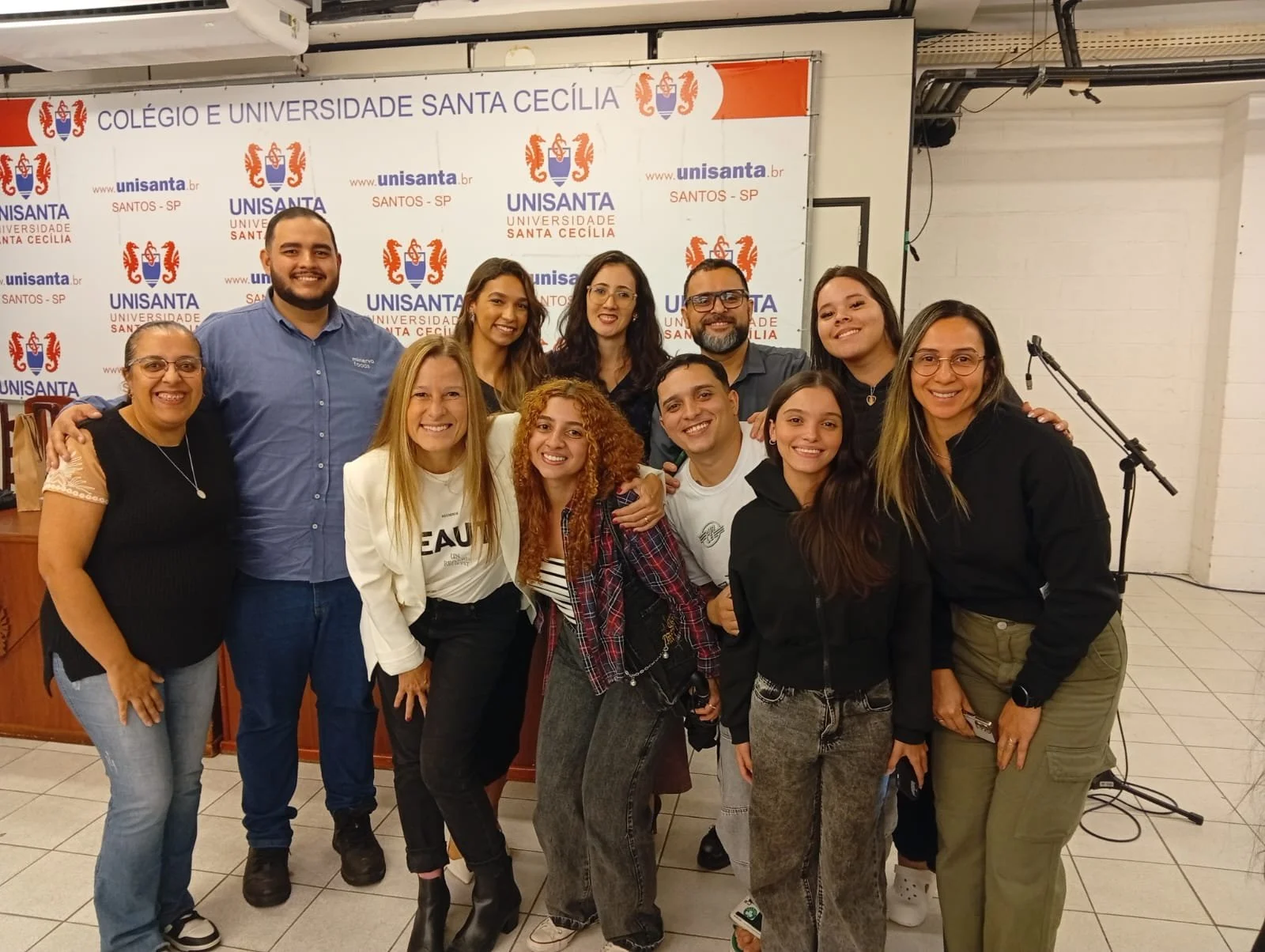 Grupo de pessoas sorrindo posando para foto em frente a uma parede com logotipo da Universidade Santa Cecília em Santos, SP, Brasil, durante evento ou reunião acadêmica.