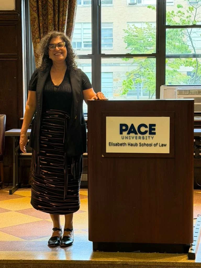 Mulher sorridente de cabelos cacheados, usando óculos, vestindo roupas pretas, posando ao lado de um púlpito com o letreiro "PACE University Elisabeth Haub School of Law" em uma sala com janelas grandes e vista para árvores e edifícios exteriores.