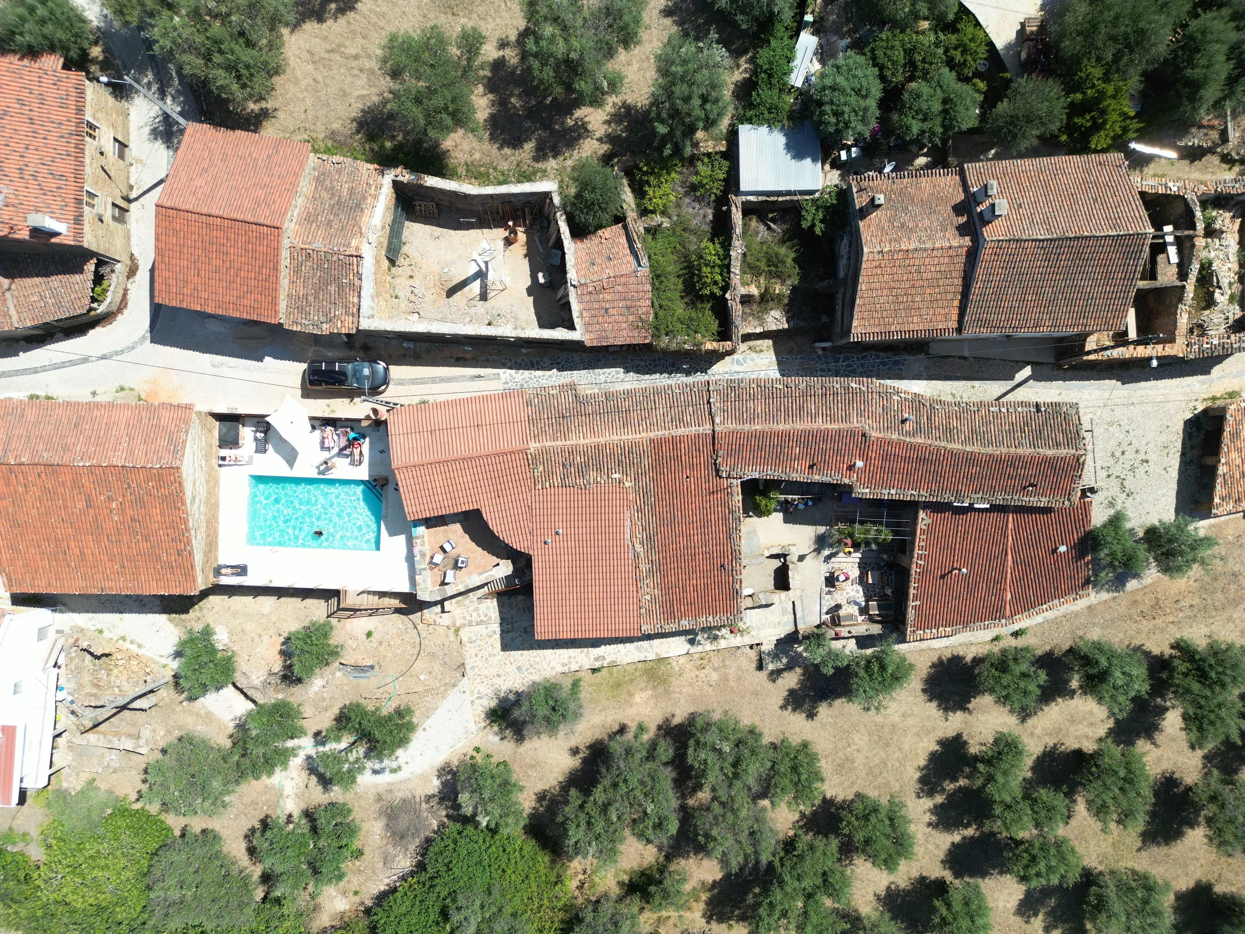 Aerial view of a ruined medieval village street in Portugal. Buildings have red-tiled roofs and are surrounded by trees. A swimming pool sits in the courtyard of a recently repaired eco tourism guesthouse amongst the disused surrounding buildings.