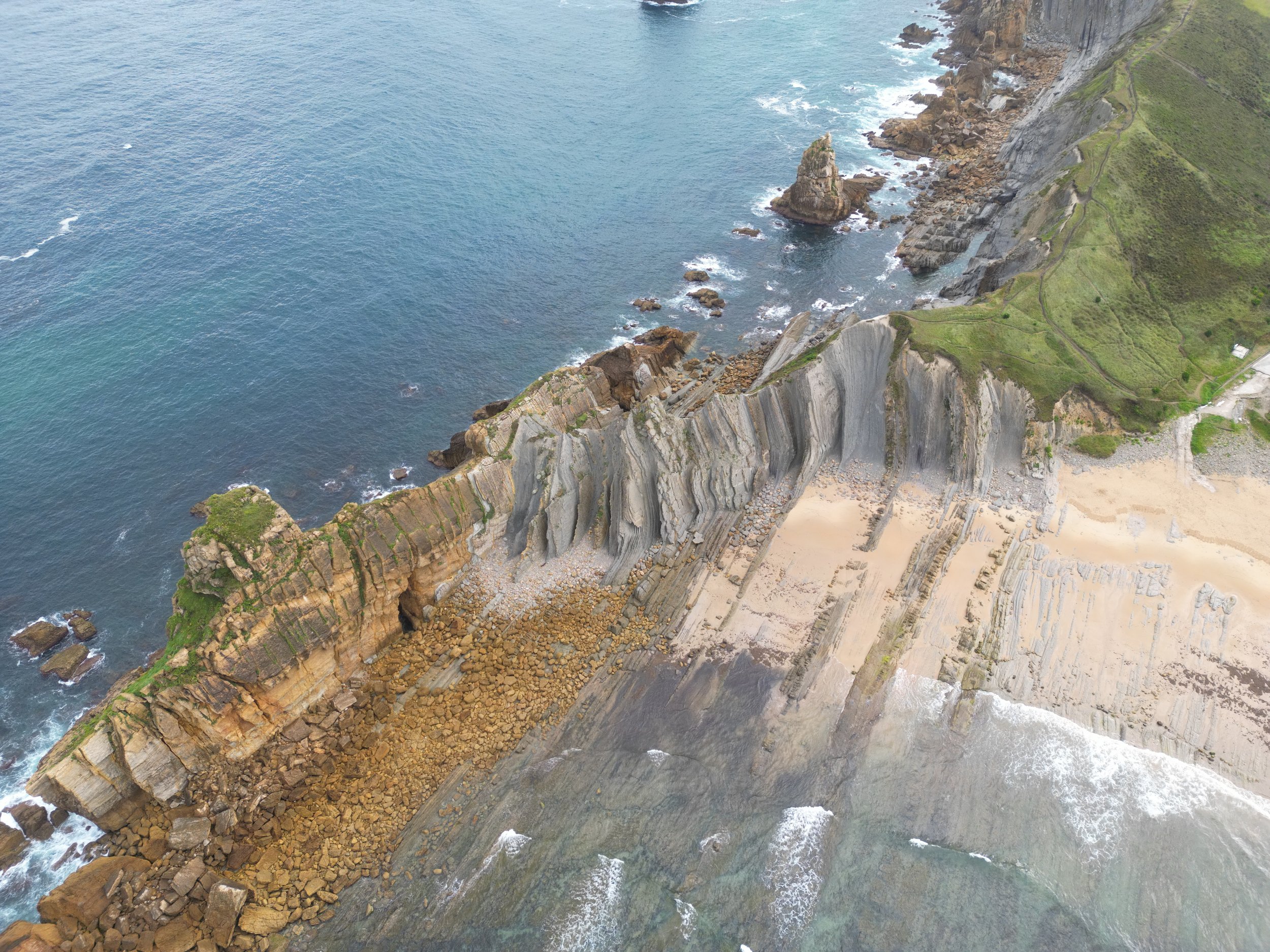 Aerial view of a rugged coastline in Northern Spain with steep cliffs, rocky formations, and a sandy beach below, adjacent to an expansive ocean.
