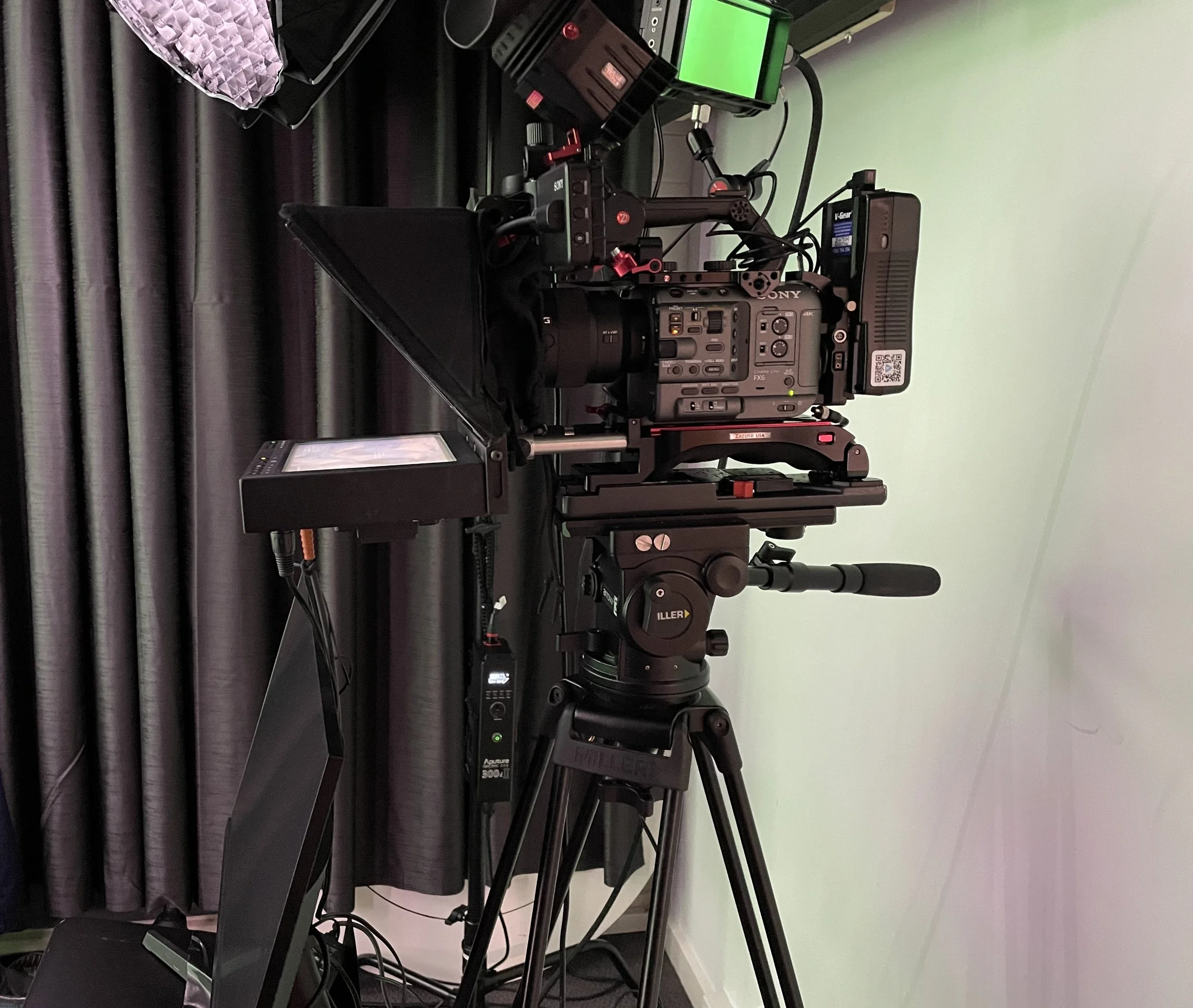 Professional video camera on a tripod set up in a studio with a light green wall and black curtains in the background.