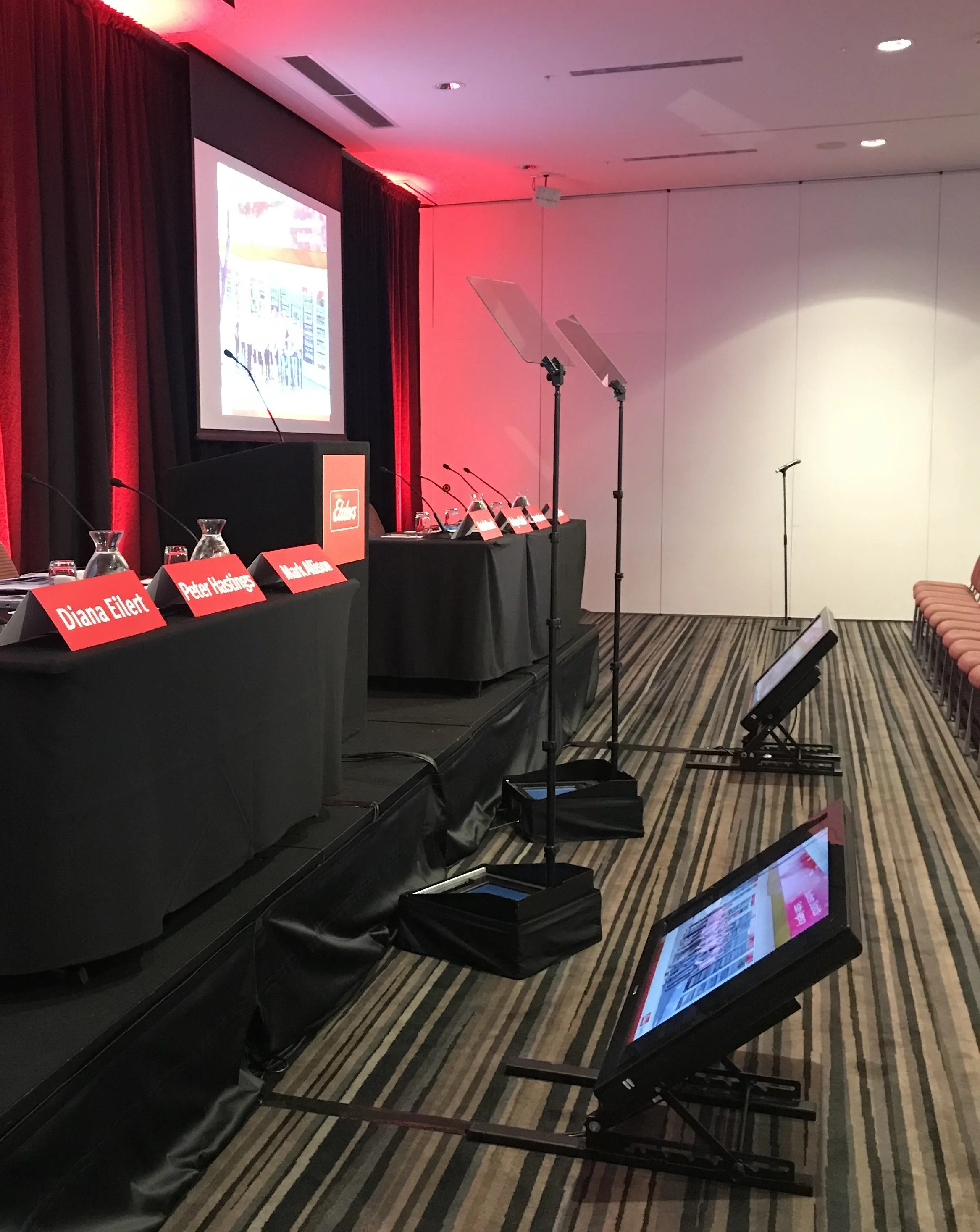 Conference room setup with a stage, table with microphones and red nameplates, two floor-standing teleprompters, projection screen, and empty chairs on the right. The room has red lighting on the walls and a striped carpet.