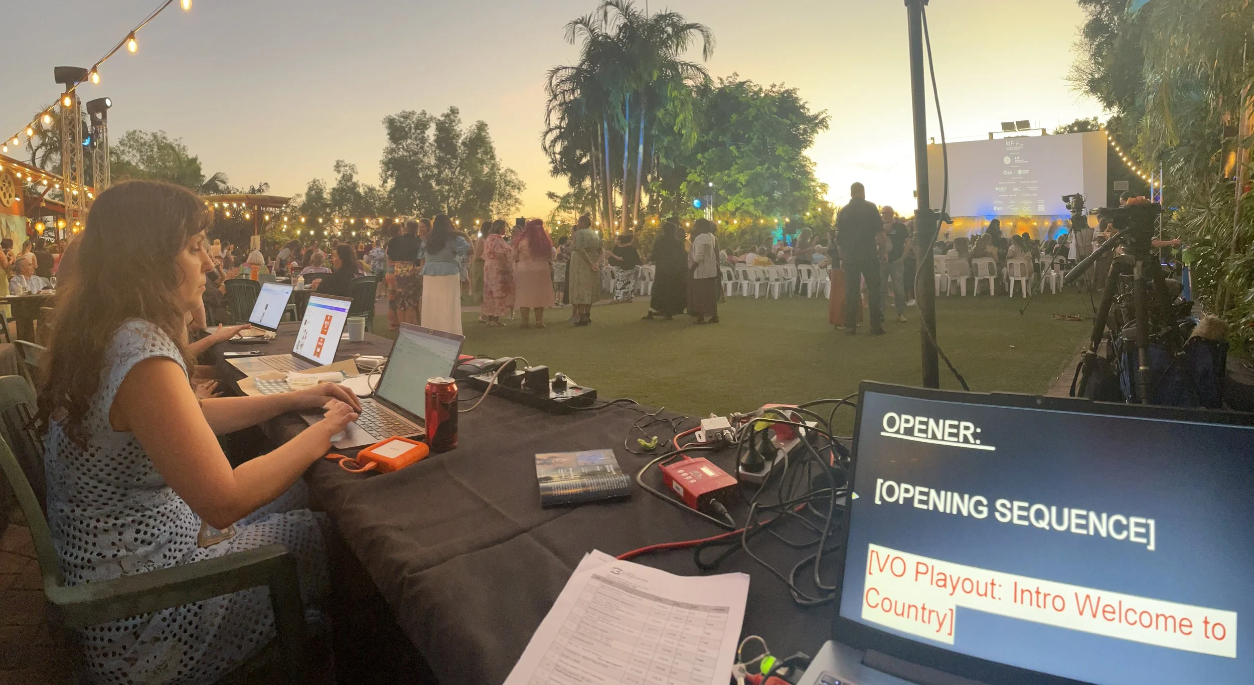 Event organizers working at a table with laptops at an outdoor event during sunset, with a large crowd gathered in front of a stage and a giant screen showing the event schedule.