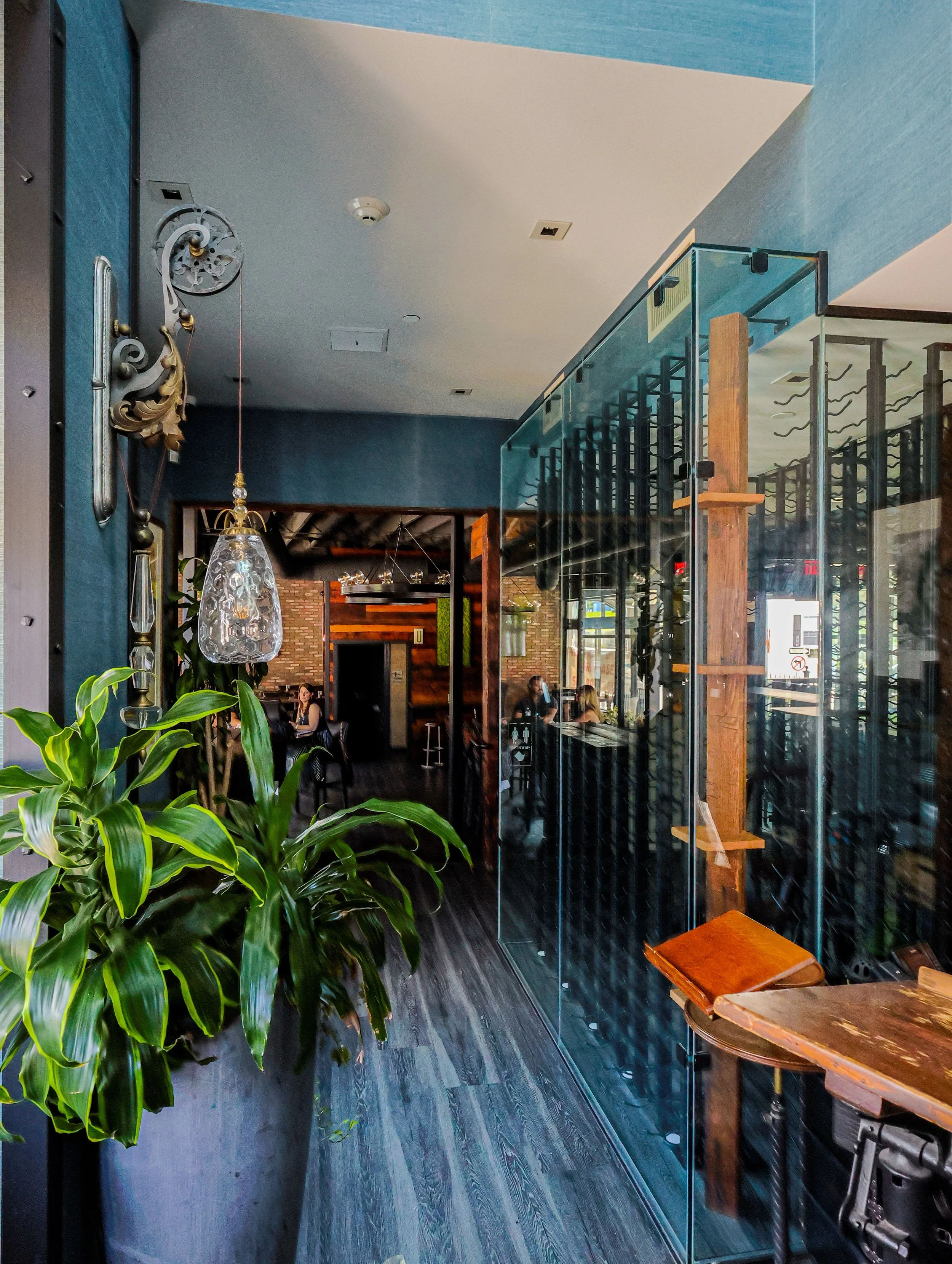 Interior of a modern restaurant with a large potted plant, glass partition, wooden accents, and patrons dining inside.