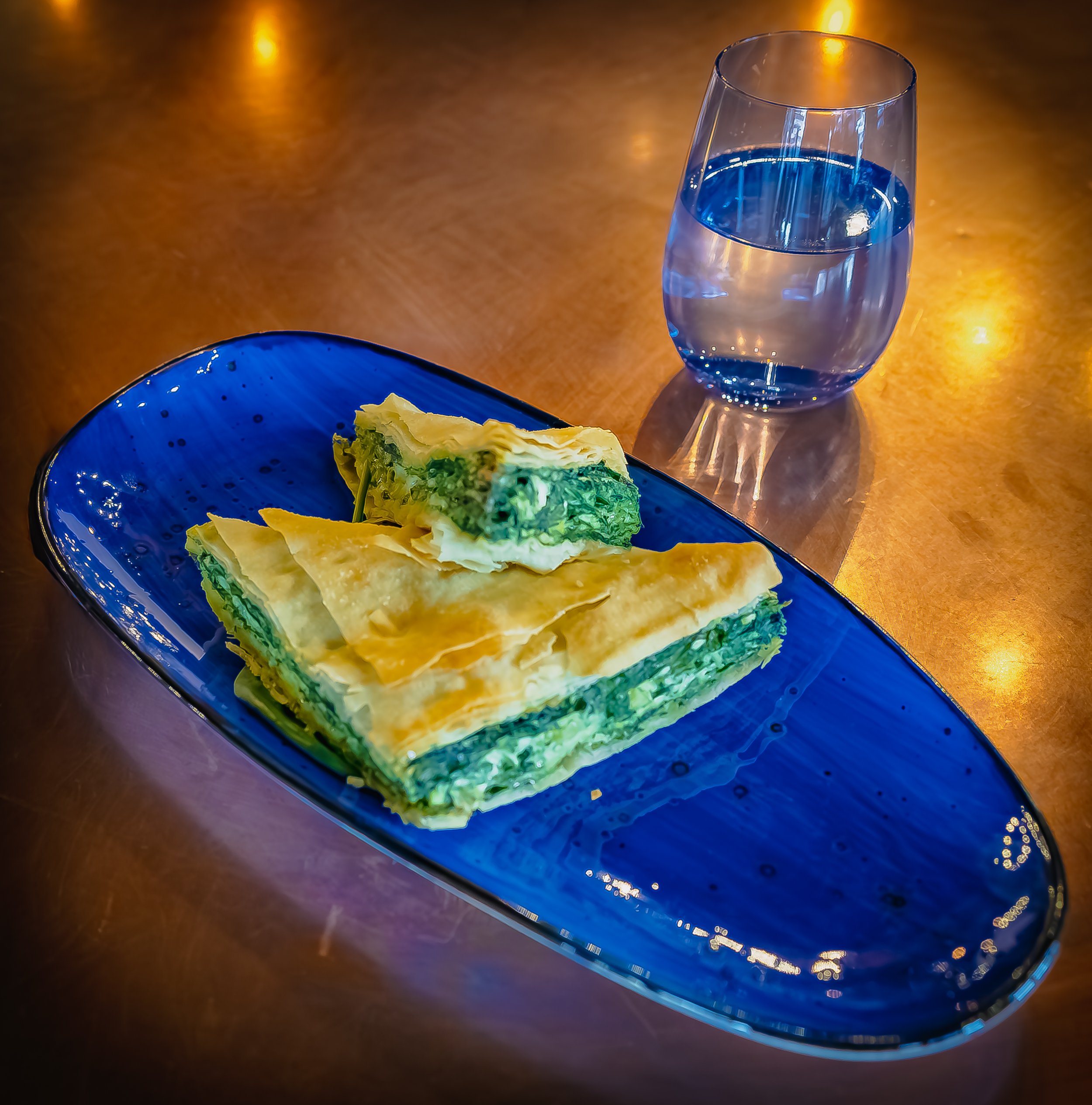 A serving of spinach and feta pastry on a blue plate with a glass of water on a wooden table.