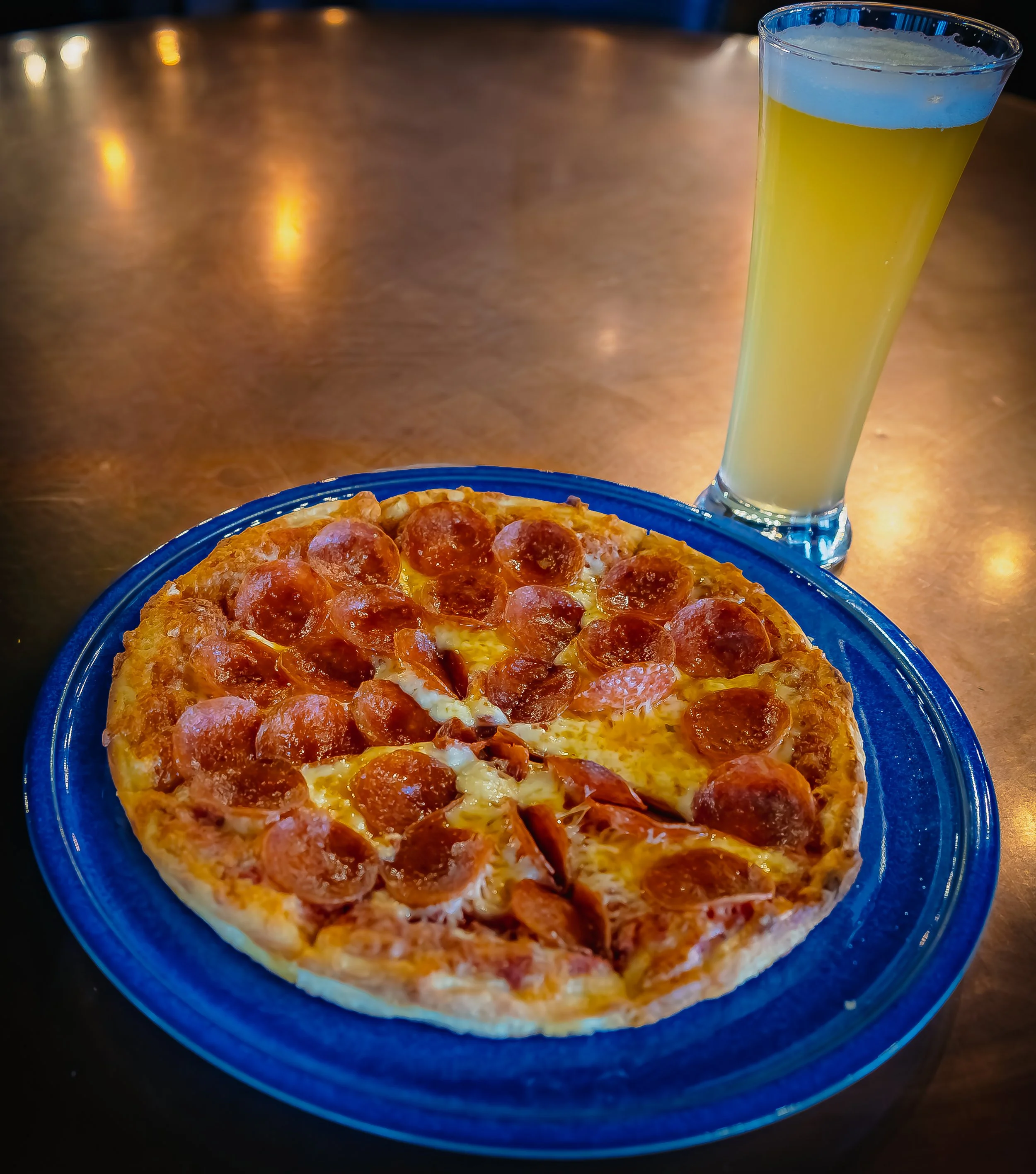 Pepperoni pizza on a blue plate with a glass of light-colored beer on a wooden table.