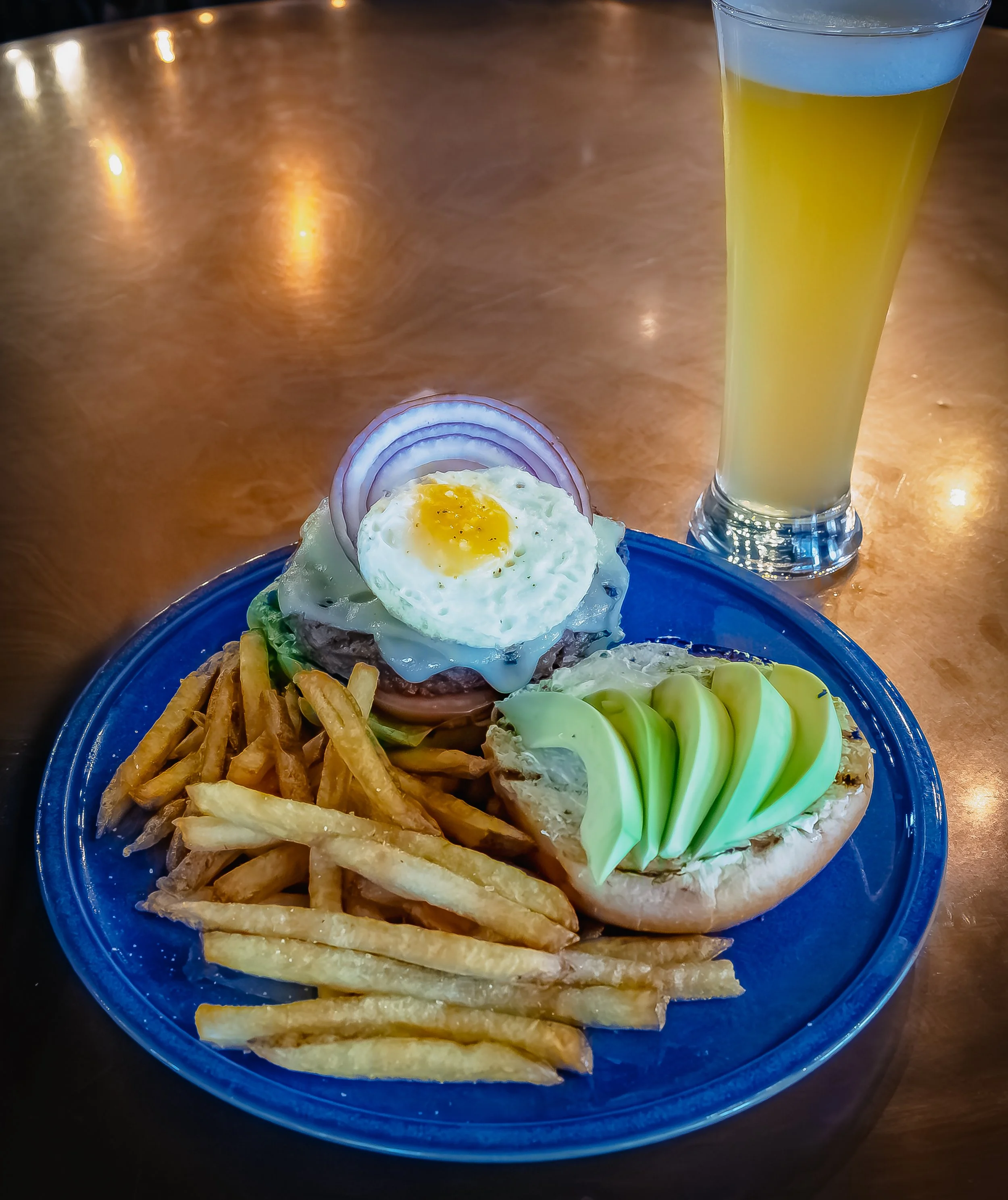 A plate with a cheeseburger topped with a fried egg and onion, a side of French fries, and a slice of bread with avocado slices, with a pale yellow drink in a tall glass in the background.
