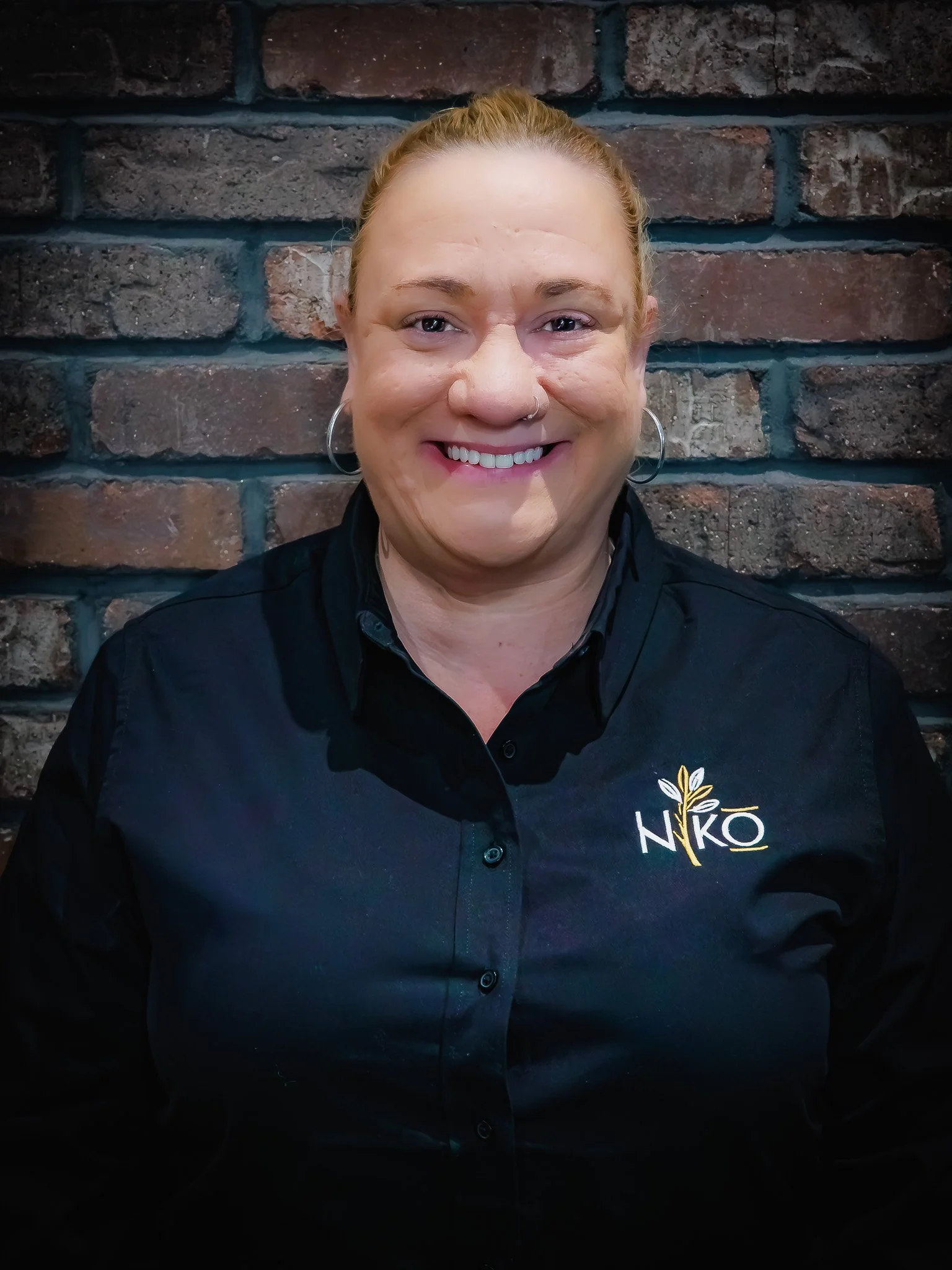 A woman with red hair in a bun, wearing a black shirt with the logo 'NIKO' on it, smiling in front of a brick wall.