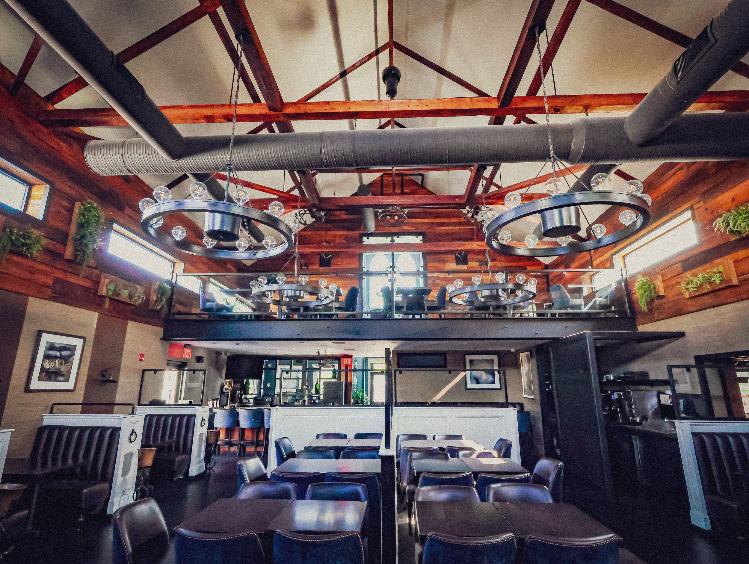 Interior of a modern restaurant with dark dining tables and chairs, a balcony with additional seating, wooden wall accents, large windows, and trendy chandeliers.