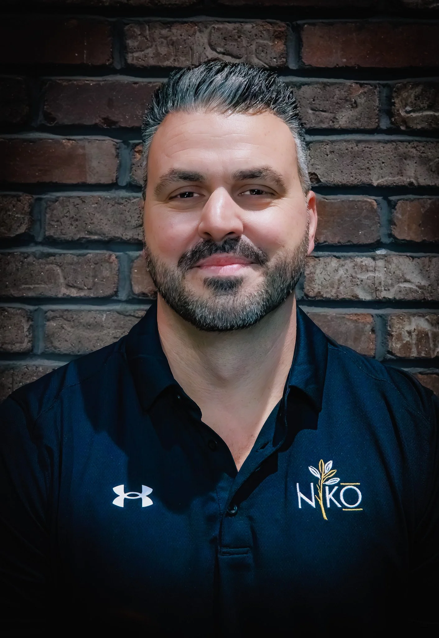 A man with short dark hair and a beard, wearing a black Under Armour shirt with a logo that says NIKO, standing in front of a brick wall.