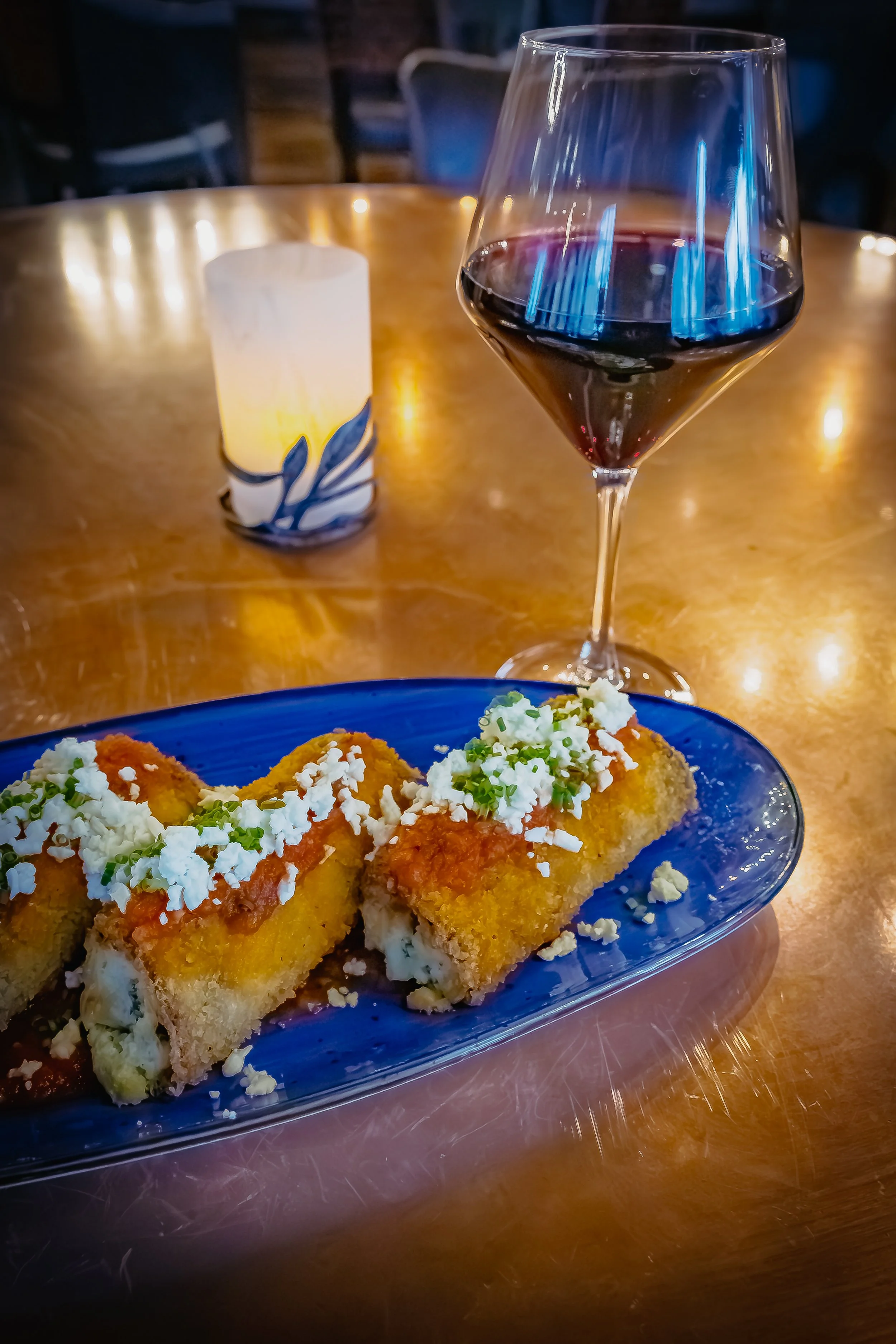 A plate of three fried appetizers topped with cheese, served with a glass of red wine. A candle in a decorative holder is also on the table.