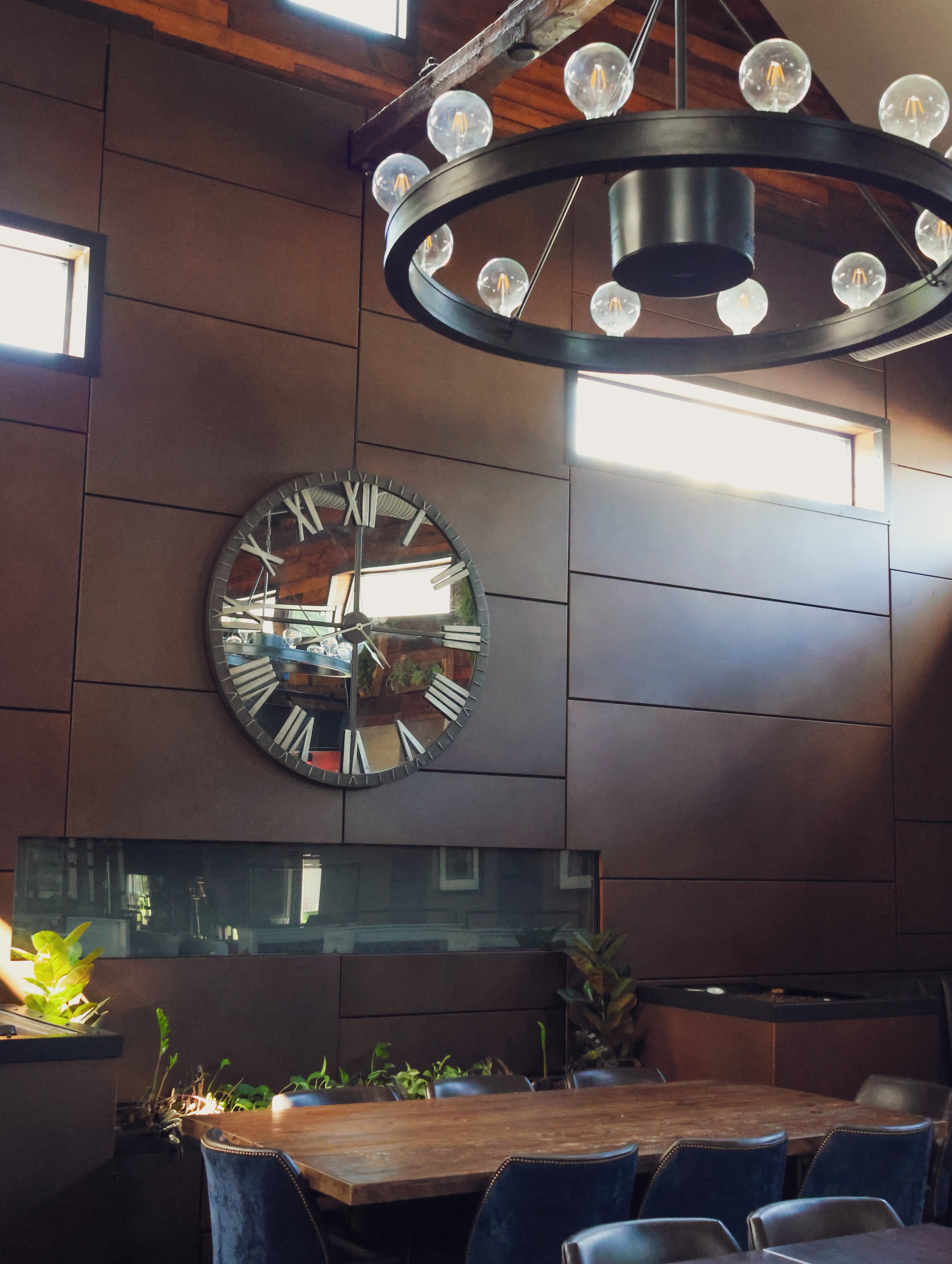 Interior of a modern dining area with a large round iron clock on a brown paneled wall, a black circular chandelier with exposed bulbs hanging from a wooden ceiling, a wooden table with dark chairs, and small indoor plants.