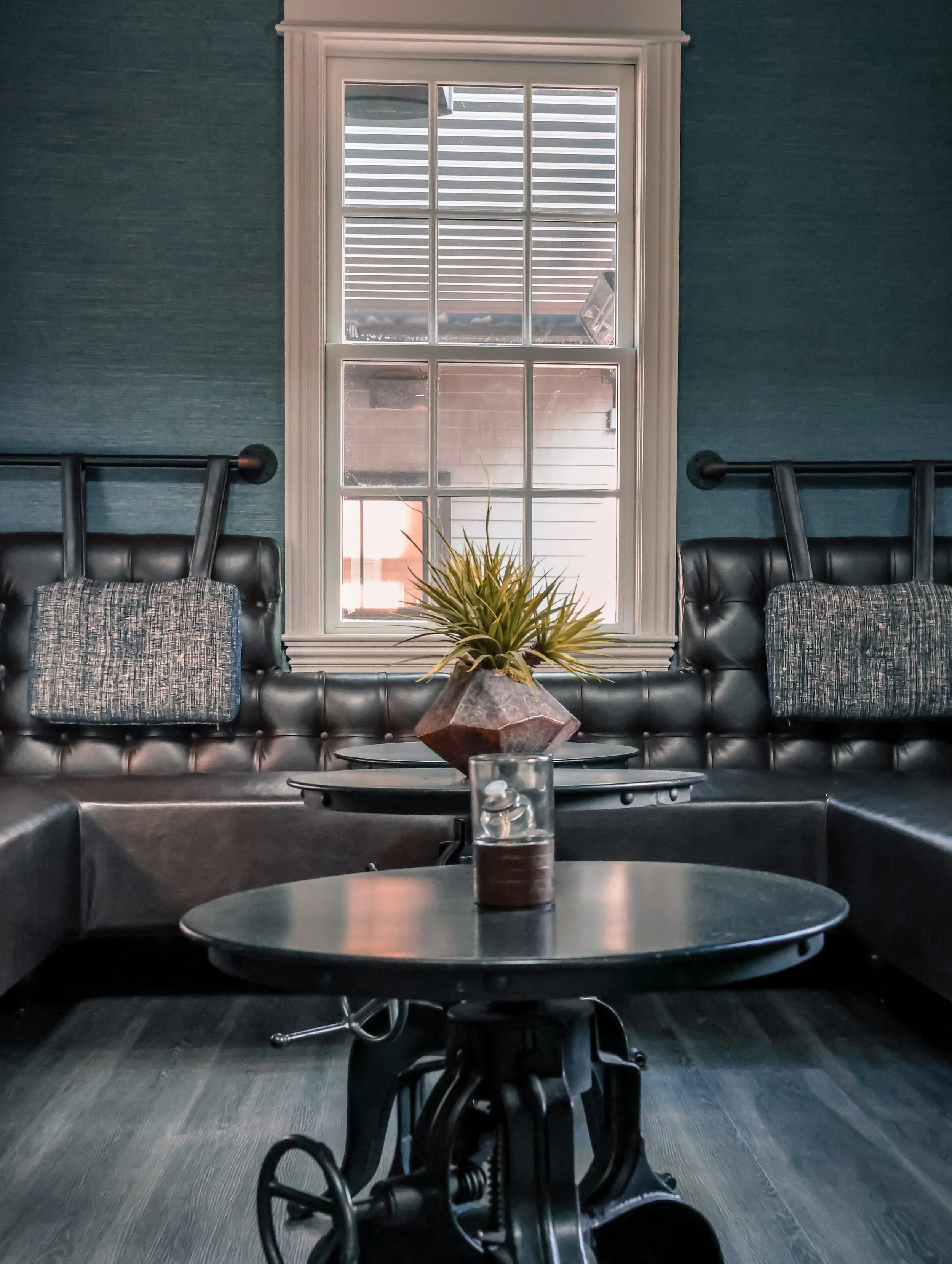 Interior of a living room with black leather sectional sofa, a window with white trim, a potted plant on a black coffee table, and dark wood flooring.