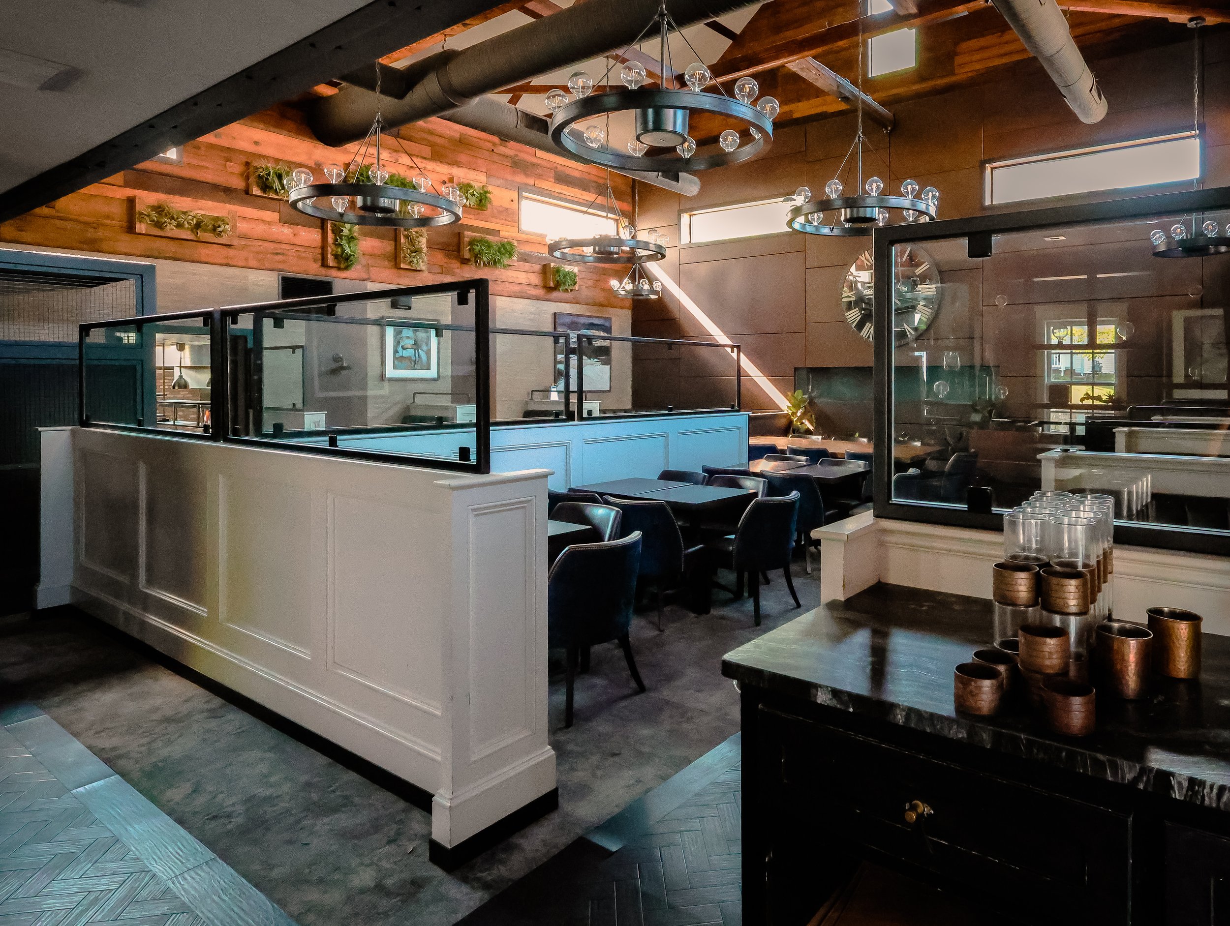 Interior of a modern restaurant featuring wooden wall panels, black chairs, and tables separated by glass partitions. Decorative ceiling lights and greenery on the wall enhance the cozy ambiance.