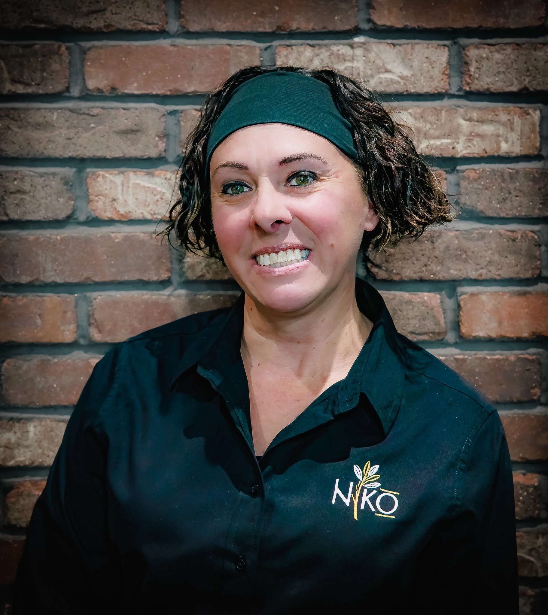 A woman with curly dark hair wearing a black headband and black shirt with NIKO logo, standing against a brick wall.