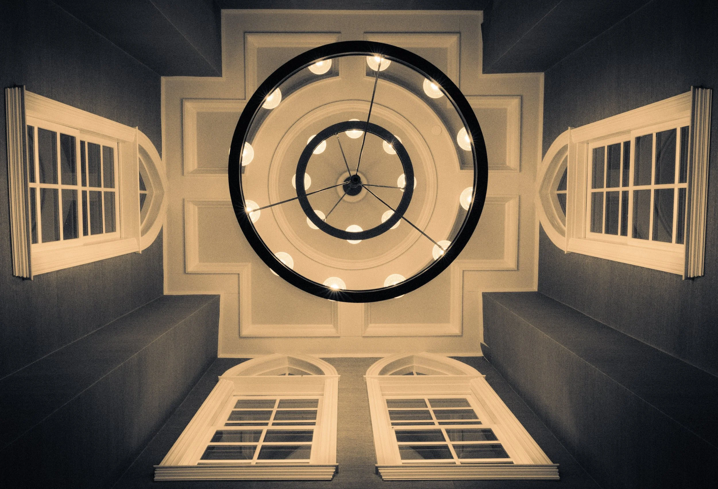 Looking up at a circular chandelier with lights, framed by four tall windows and decorative molding in a house interior.